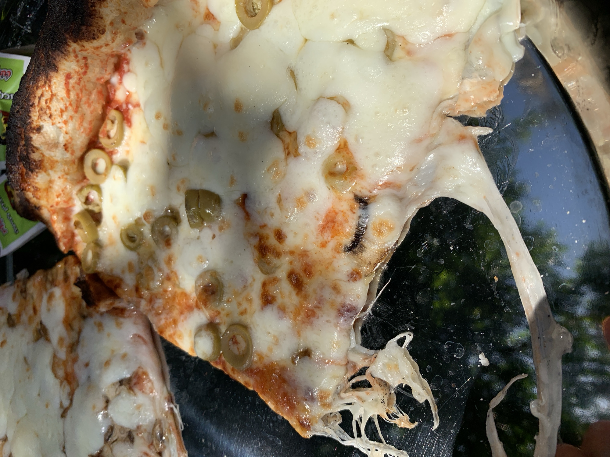 A high-angle shot depicts a pizza slice, overflowing with melted cheese and green olives, on a black, possibly glass, surface. The crust is charred in areas, and visible under the cheese is red sauce. Part of another pizza slice sits at the bottom-left corner of the image. Based on the presence of a green food product packaging featuring Hebrew characters and the background suggesting an outdoor location, this scene may be in Tel-Aviv, Israel. The lighting is bright and casts shadows.