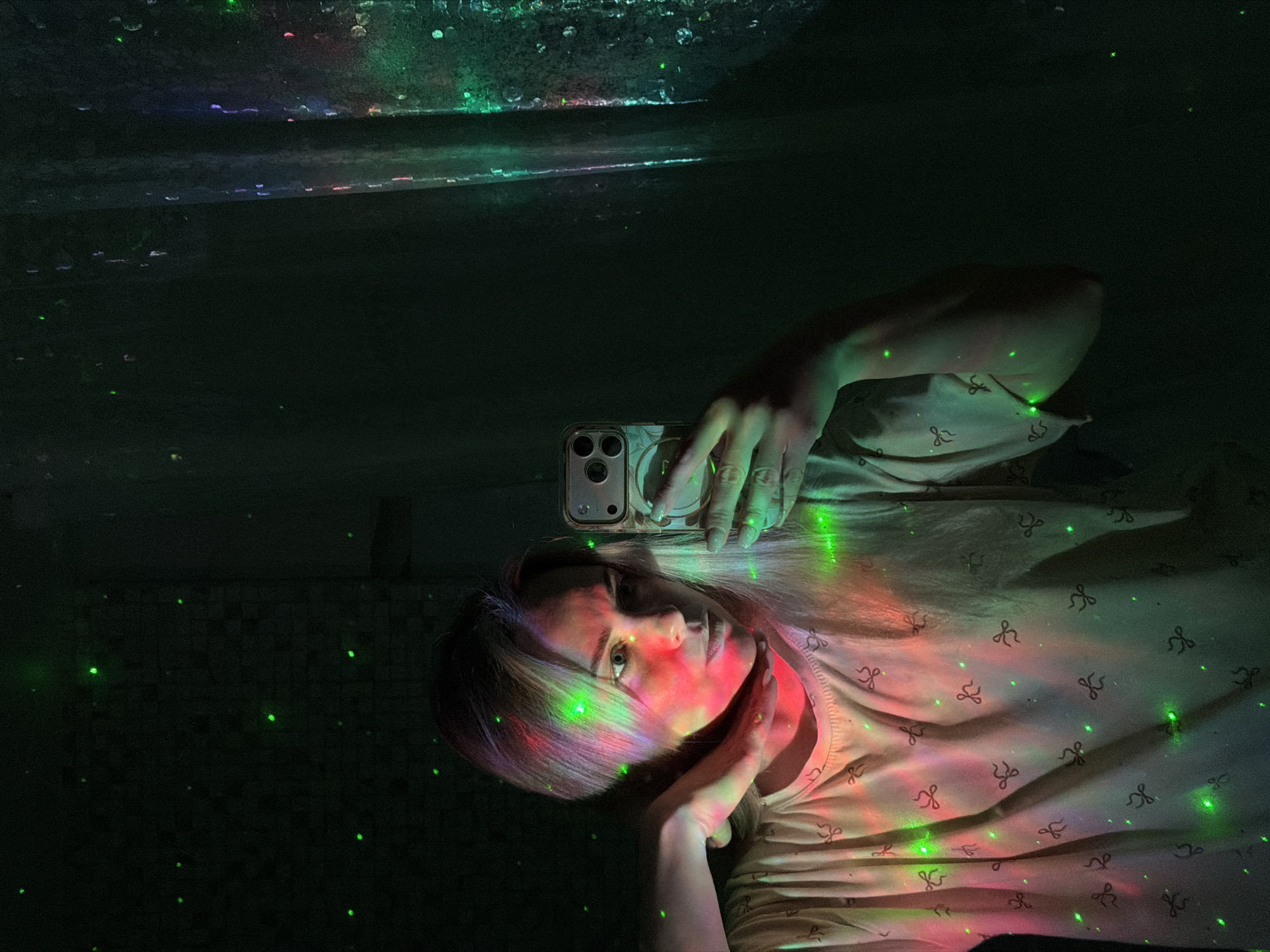 The image displays a selfie portrait of a young woman captured in a dimly lit indoor environment in Votkinsk, Russia, dramatically illuminated by colorful light projections.
The woman, with fair skin and light-colored hair, is positioned with her head slightly tilted, looking directly towards the camera. Her right hand holds a modern smartphone with three visible camera lenses above her head, indicating she is taking the photograph. Her left hand gently rests on her jawline. She is wearing a light-colored top, possibly a t-shirt, adorned with a repeating pattern of small, dark bow-like shapes.
The scene is strikingly lit by a projector that casts dynamic red, green, and possibly purple light beams across her face, hair, and clothing, creating vibrant streaks and shadows. Numerous small, bright green laser-like dots are scattered throughout the frame, appearing on her body, clothing, phone, and the dark background. This creates a lively and atmospheric mood. The strong contrast between the bright, colorful lights and the dark surroundings suggests a nighttime setting or a very dark room.
The background is predominantly dark, with some faint textures and subtle reflections visible in the upper section, possibly a ceiling or wall. The woman maintains a neutral and calm expression. No discernible text is visible on her clothing or the phone.