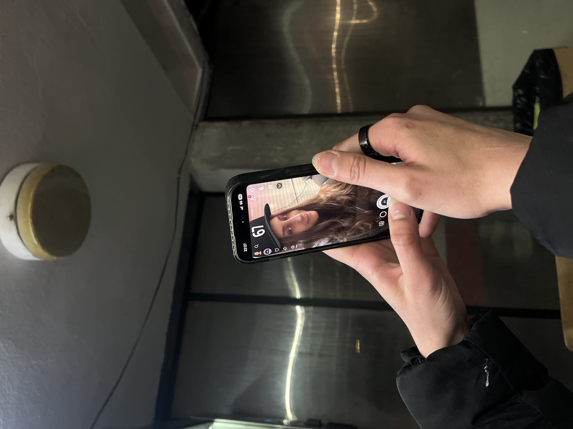 A person wearing a black ring and a black jacket is holding a black smartphone with a camera app open. The phone screen displays a selfie of a woman. The background shows a light fixture on a white wall, a gray surface, and a black vertical bar. No details can be confirmed that determine the location of Lozarevo, Bulgaria.