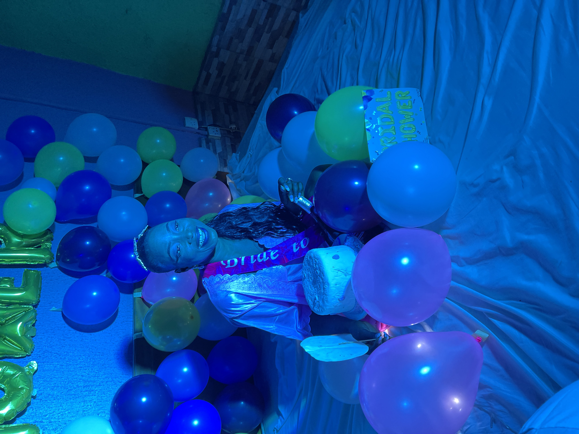 A person lies amidst numerous balloons of various colors, including blue, yellow, and pink. The person wears a "Bride to Be" sash and a tiara. There is a "Bridal Shower" sign present. The scene is lit with blue light. The location is Akwanga, Nigeria.