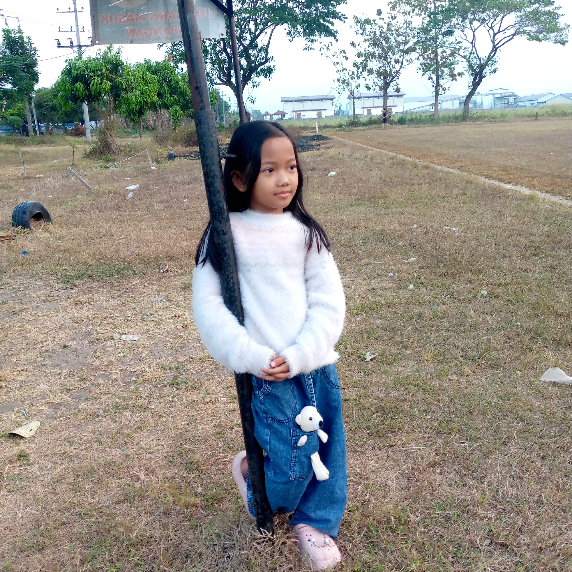 A young girl with long, dark hair stands next to a dark pole, leaning on it with her left arm. She is wearing a white fluffy sweater and blue jeans. A small white stuffed toy is attached to her jeans pocket. She is looking to her right, with her hands clasped in front of her. The background is an outdoor scene with dry grass, some trees, and buildings in the distance. The location is identified as Pacing, Indonesia. The scene appears to be a park or an open field. There is a discarded tire on the left side of the image. The sky is overcast.