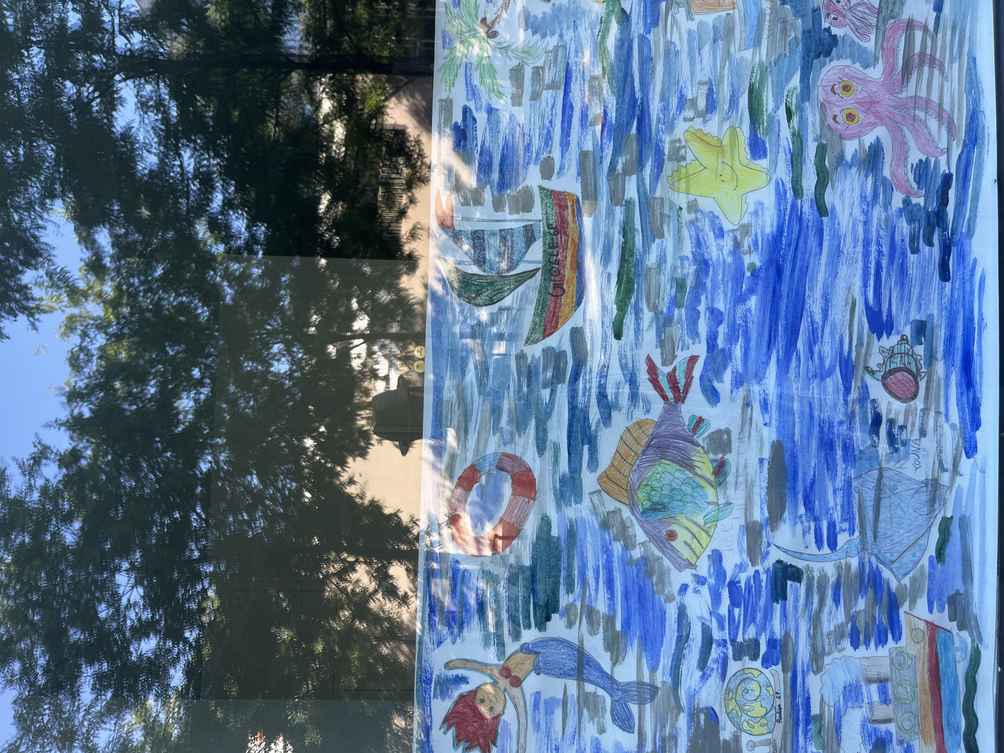 This is a photograph of a children's drawing displayed behind a reflective surface, likely a window. The drawing depicts an underwater scene filled with various marine life and objects. In the upper left corner, a mermaid with red hair and a blue tail is swimming. To her right, a life preserver floats in the water. A sailboat with green and orange sails is visible in the upper center, with the name "GIOCLEGA" written on its side. Further to the right, a palm tree suggests an island or coastal element.
The underwater environment is rendered with broad strokes of blue paint, representing water. Several fish are depicted, including a large purple and yellow fish with green accents. A snail on a small raft is in the lower left, followed by a steamship. A blue stingray is swimming in the lower center of the drawing, and a yellow starfish with a smiley face is positioned in the upper right quadrant of the underwater scene. In the lower right, a pink octopus with yellow eyes and two smaller octopuses are shown.
The reflection on the surface shows the silhouettes of trees and foliage against a bright blue sky, indicating an outdoor setting. The reflection also reveals a glimpse of what appears to be a building and possibly a hat on a chair in the background. The context of Geneva, Switzerland, is not discernible from the visual elements of the image itself.