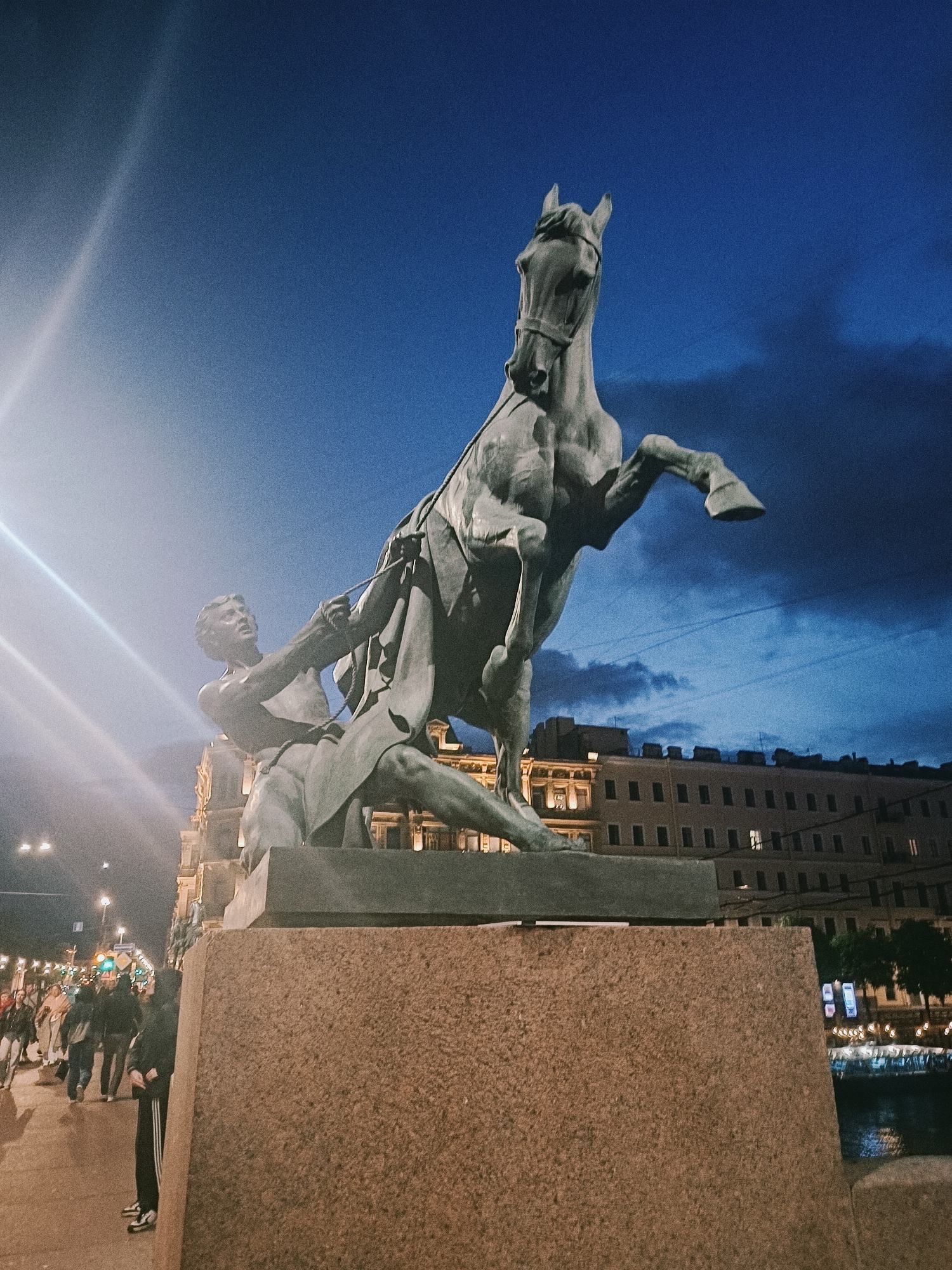 A nighttime shot captures the Anichkov Bridge horse tamers sculpture in Saint Petersburg, Russia. The bronze statue depicts a muscular man struggling to control a rearing horse. The sky is a deep twilight blue with some cloud cover, and the buildings along the embankment are illuminated, casting a warm glow. People are walking along the sidewalk adjacent to the bridge. A stone plinth supports the statue. The Neva River is visible in the background.