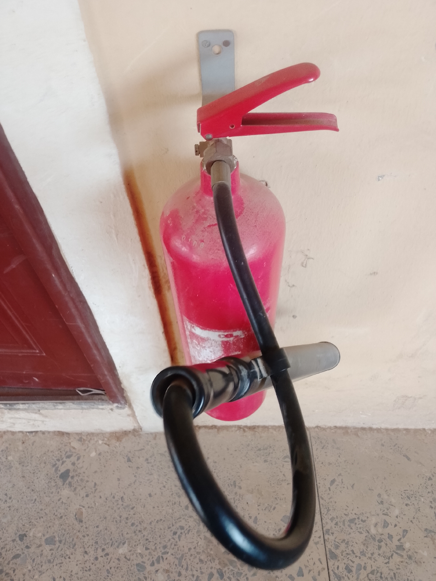 The image shows a red fire extinguisher mounted on a beige wall. The extinguisher has a black hose attached to its nozzle, which is extended downwards. The handle of the extinguisher is red and appears to be made of plastic. The extinguisher is covered in a fine layer of dust, suggesting it may not have been used recently.
The scene is set indoors or in a covered outdoor area, as indicated by the wall texture and the presence of a dark red door frame on the left side of the image. The floor is a terrazzo-like material with visible pebbles.
There is no specific activity or event taking place in the image; it appears to be a static depiction of the fire extinguisher. The lighting suggests it could be daytime, but there are no strong indicators of the time of day or weather conditions. The overall impression is one of a functional safety device that is present in its environment, possibly in a building or hallway.
No discernible text is visible on the fire extinguisher or its surroundings. The focus of the image is the fire extinguisher itself and its immediate environment.