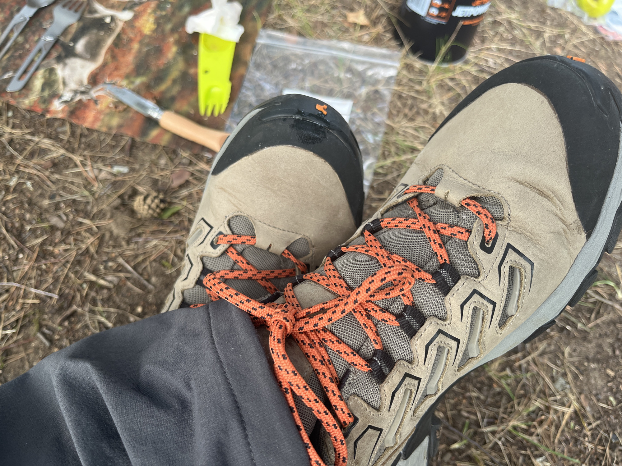 The image shows a close-up view of a person's feet wearing hiking boots with bright orange laces, standing on a forest floor. The boots are tan with black accents and appear to be well-worn. Dark gray hiking pants are visible on the left side of the frame. In the background, there is a camouflaged mat with camping cutlery, including a knife and fork, and a small white object, possibly a napkin or tissue. A clear plastic bag and a bright yellow utensil are also present. The ground is covered with pine needles and dry grass. The scene is likely outdoors in a natural environment. There is no visible indication of the specific location within Pillapalu, Estonia.