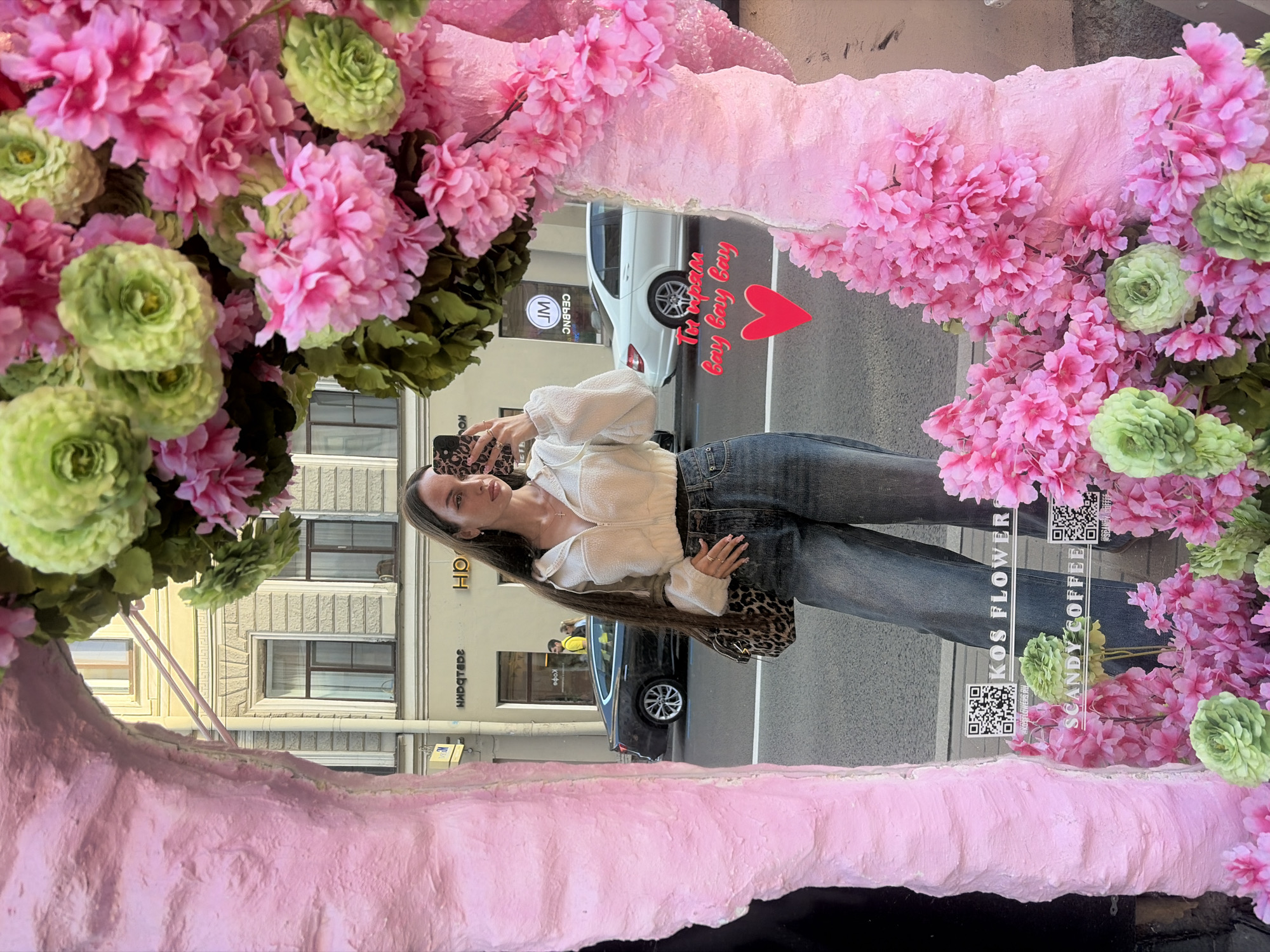 A young woman with long dark hair, wearing a cream-colored zip-up hoodie and blue jeans, stands in front of a shop window in Saint Petersburg, Russia. She is taking a mirror selfie with her phone, which is held up to her face. The shop window is decorated with a large, bright pink, textured sculpture that is adorned with numerous pink and green artificial flowers. Behind the woman, the reflection in the window shows a street scene with buildings, cars, and people. One of the buildings has signage that includes the words "KOS FLOWERS" and "SCANDY COFFEE," indicating the location. A red heart and text overlaying the reflection read "Ты играй Bay Bay Bay". The overall scene is vibrant and colorful, with a focus on the decorative elements in front of the shop.