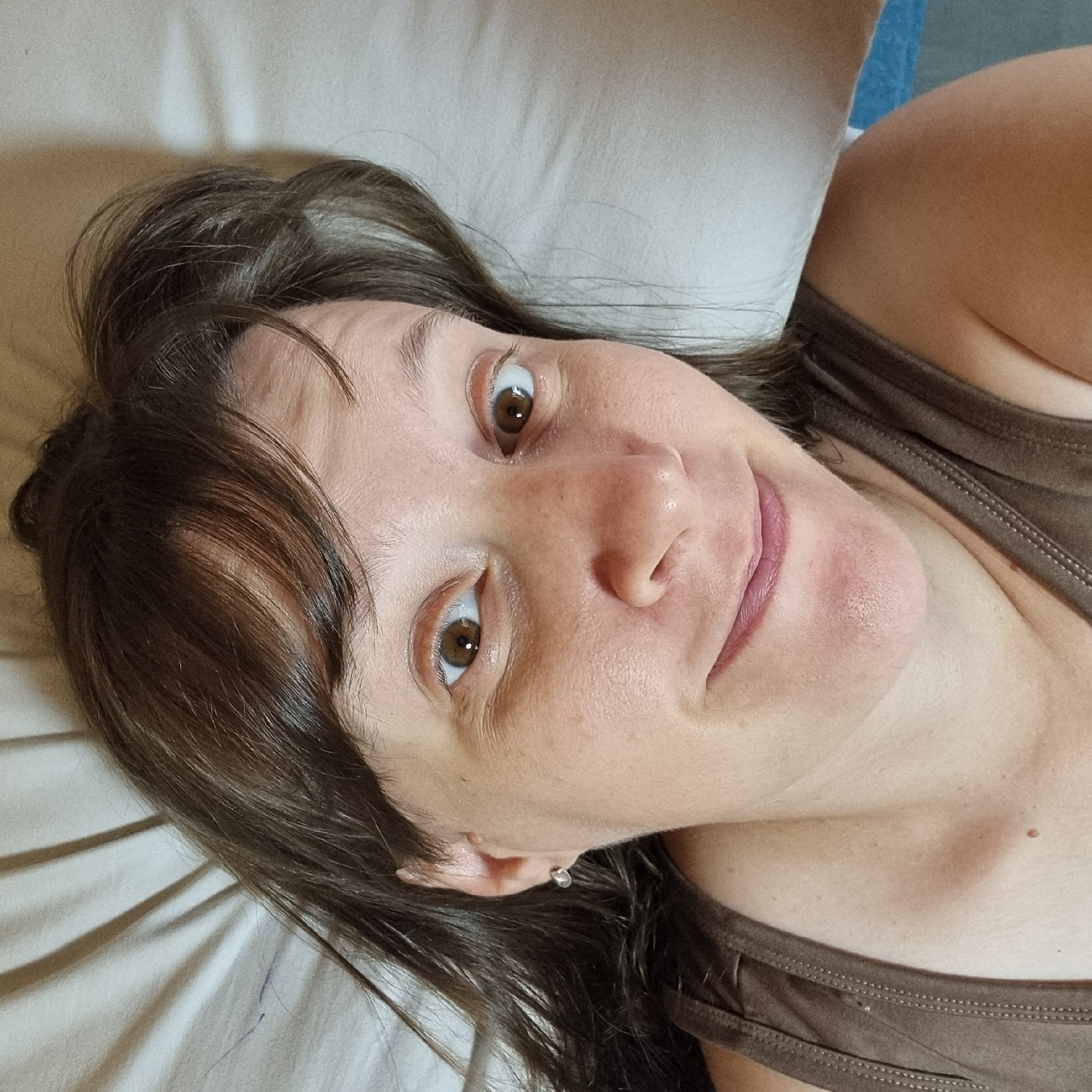 An overhead shot shows a woman lying on her back on a white surface, possibly a bed. She has brown hair that is falling around her face and shoulders. She is wearing a brown tank top. The woman is looking directly at the camera with a slight smile on her face. Her eyes are wide open and her pupils are dark brown. There are some shadows on the white surface behind her, suggesting a light source is present. The overall impression is of a casual, candid photograph. The location is not specified in Dunnae-myeon, South Korea.