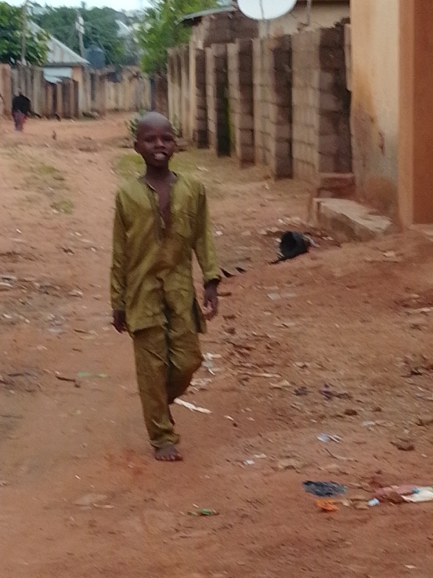 A young boy walks down a dirt road in a Nigerian city. He is wearing a loose-fitting, olive-green outfit. The road is sandy with scattered debris. Buildings with corrugated metal roofs and brick walls line the street, some appearing to be in disrepair. In the background, other figures and structures are visible, suggesting a residential area. The sky is overcast.