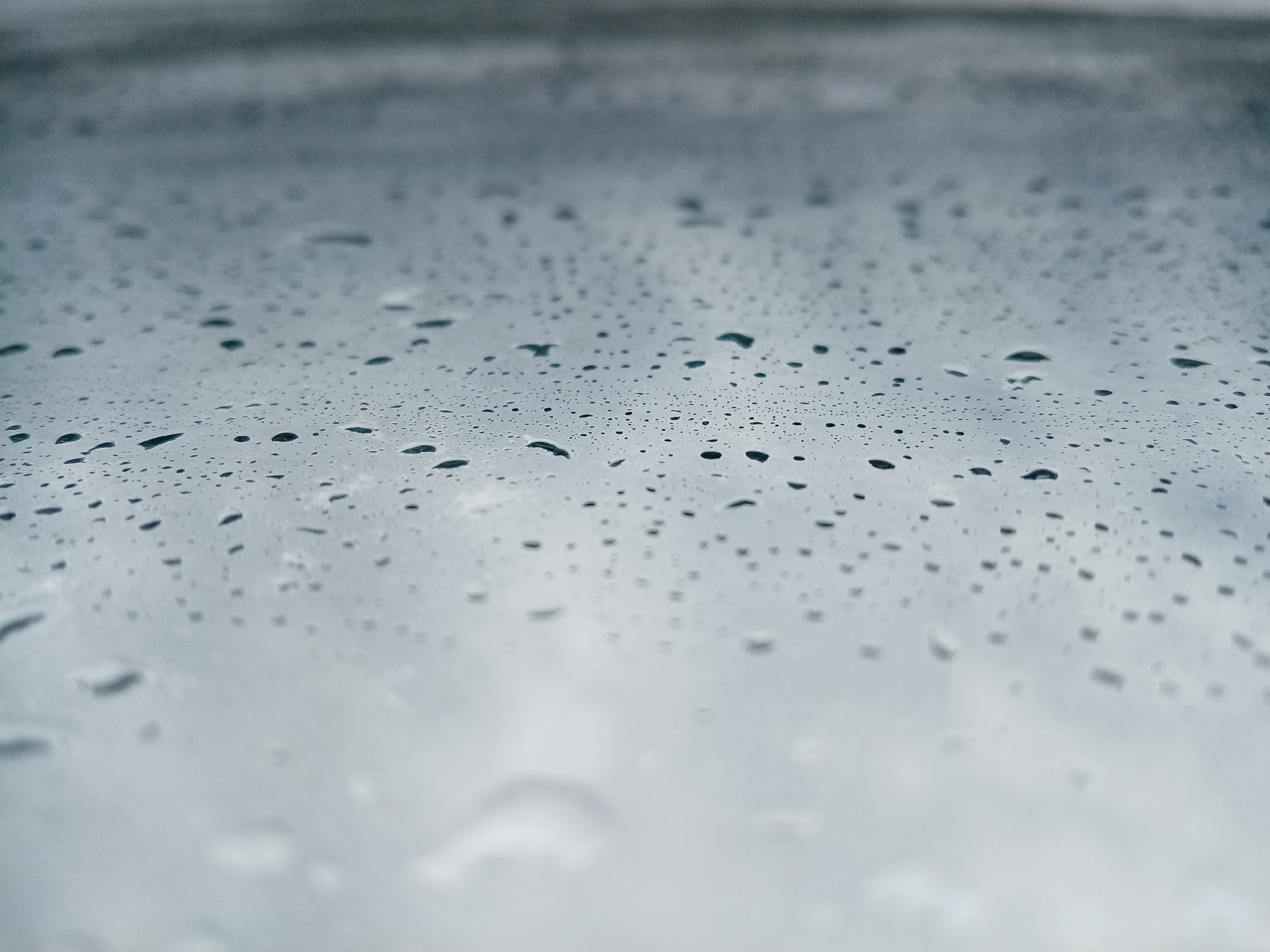 The image shows a close-up view of raindrops on a surface, possibly glass. The background appears to be a blurred cityscape, suggesting an urban environment, or a cloudy sky. There is no indication in the image that points to the specific location of Izhevsk, Russia. There are no people, actions, or interactions visible in the scene.