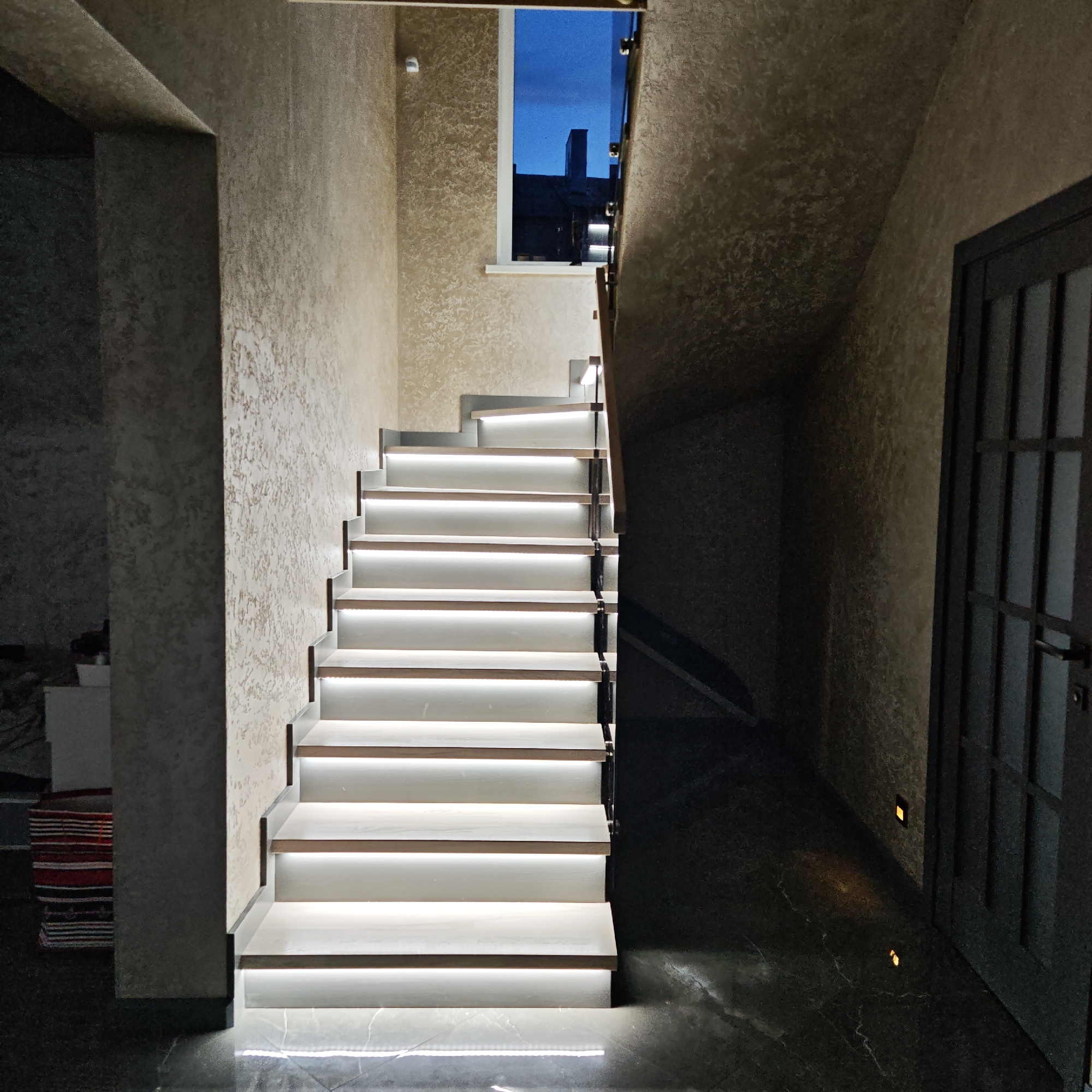 The image depicts an interior scene featuring a staircase with integrated lighting. The steps are primarily white with a wood trim, and LED lights are installed underneath each step, illuminating the treads and the surrounding floor. The staircase ascends towards a small window, through which a dark, possibly nighttime, sky and rooftops are visible. The walls have a textured beige finish. To the right of the staircase, a dark doorway with a multi-pane glass window is visible. The floor is dark and glossy, reflecting some of the light from the stairs and the two small lights set into the base of the wall next to the doorway. To the left of the staircase, a partial view of a room reveals some furniture and what appears to be a striped rug. The photograph was taken in the city of Rostov, Russia.