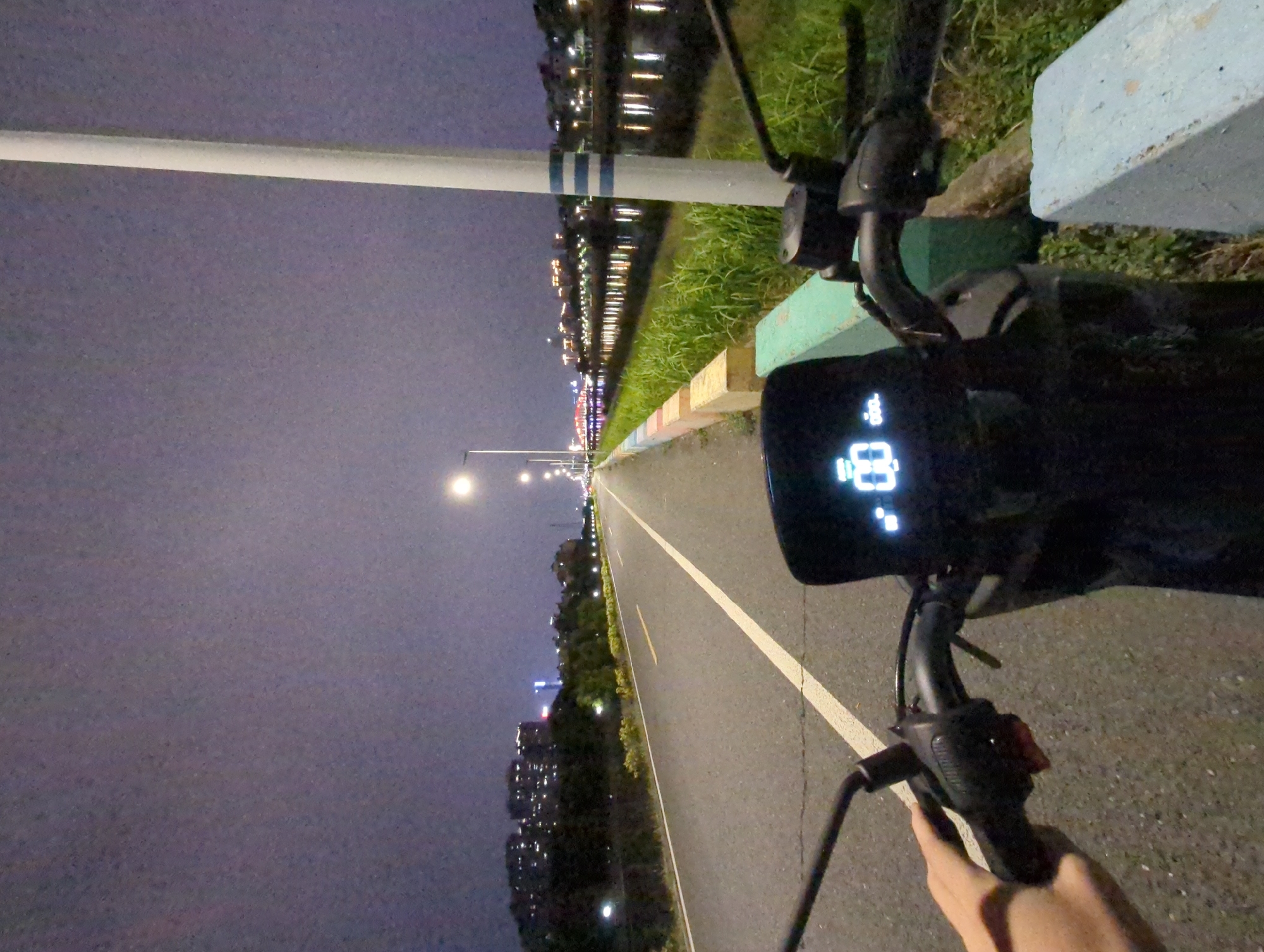 A person is riding a scooter or bicycle on a paved path with a white line marking the edge. Streetlights illuminate the path, which stretches into the distance. On the left side of the path, there are buildings and trees. On the right side, there is grass and a body of water reflecting lights from the city. The scene appears to be at night, in Shunde District, China. The scooter or bicycle's dashboard displays information, possibly including speed. The person's hand is visible near the scooter's controls.