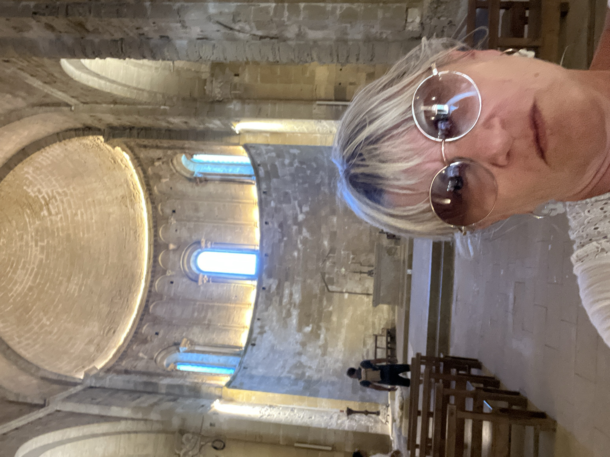 A woman with blonde hair and sunglasses takes a selfie in the interior of a historic stone church, likely the Cathédrale Saint-Jean-Baptiste de Maguelone, in Villeneuve-lès-Maguelone, France. She is positioned in the foreground, her face filling the lower portion of the frame. Behind her, the church's grand architecture is visible, featuring a large, domed ceiling, arched windows with blue-tinted glass, and stone walls. Pews are arranged in rows on the left side of the church, and an altar area is visible in the background. A man wearing a backpack is seen standing in the middle ground, facing away from the camera, towards the altar. The overall lighting suggests an interior setting with ambient light filtering through the windows and possibly some artificial lighting enhancing the dome.