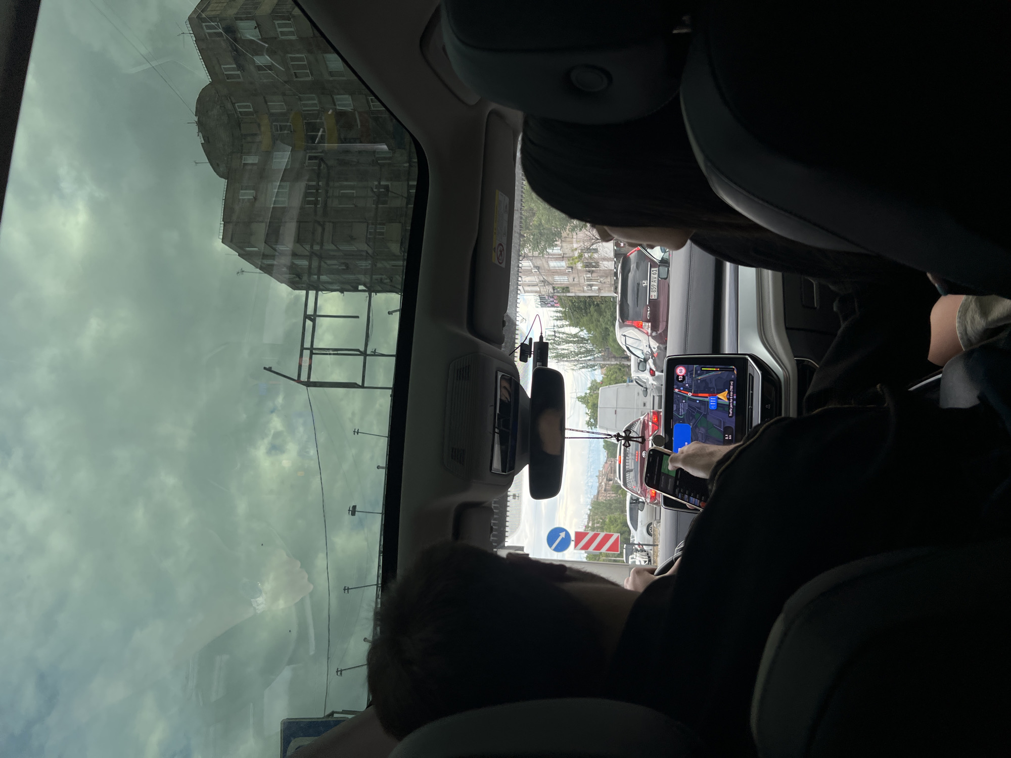 The photograph, shot from within a vehicle, captures a scene in Yerevan, Armenia. Outside the vehicle's window, a multi-story building dominates the landscape, its weathered facade and architectural details characteristic of the city. The sky is overcast, lending a muted tone to the scene. Through the windshield, urban traffic can be observed.
Inside the vehicle, the primary focus is on the driver and front passenger. The driver is seen holding a mobile phone, seemingly interacting with its interface. The car's infotainment system displays a navigation screen, indicating route guidance. Traffic conditions are also visible on this screen.