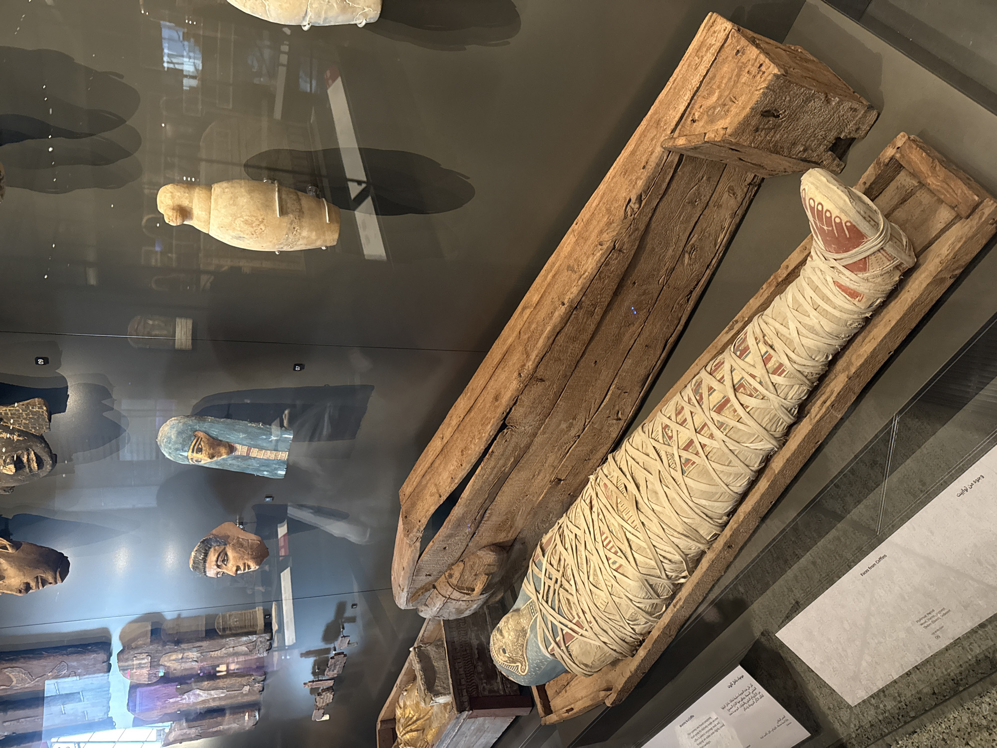 In the city of Kafrat Nassar, Egypt, an exhibition case displays ancient artifacts. The primary focus is a mummified body, wrapped in linen bandages, resting within a wooden sarcophagus. The mummy's head is adorned with a blue headdress, and its feet are painted with red and white detailing. Beside the sarcophagus lies an open, larger wooden coffin. In the background, several ancient Egyptian masks, made of wood and painted with human features, are mounted on a dark wall, some with blue headdresses. Further back, a few ceramic vessels are visible. The exhibition is presented within a museum setting, with glass cases separating the artifacts from the viewer.