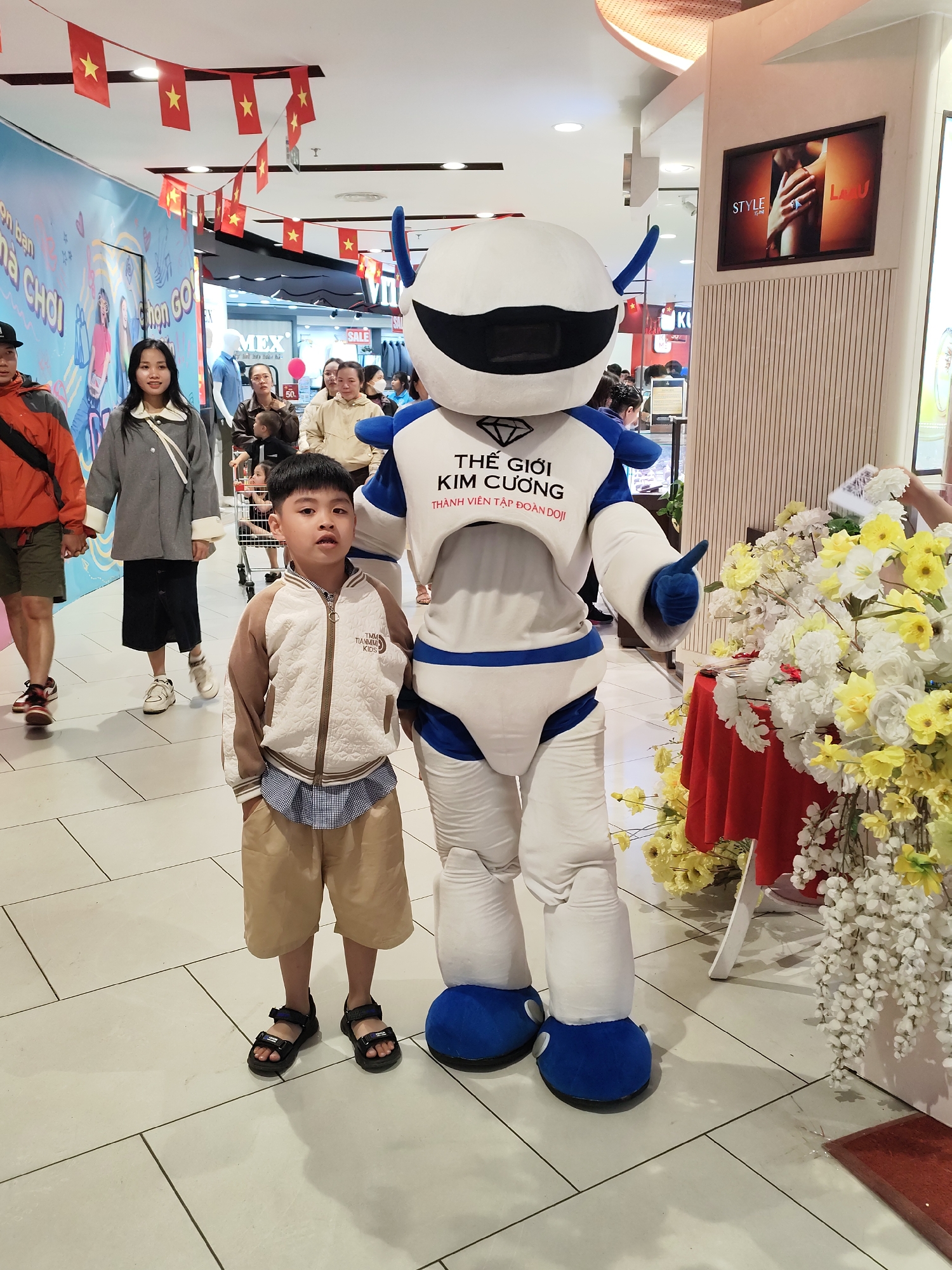A young Asian boy stands next to a robot mascot inside a brightly lit shopping mall. The boy is wearing a tan and white jacket, a blue and white checked shirt, tan shorts, and black sandals. The robot mascot is white with blue accents and has a large black visor for a face. Text in Vietnamese is visible on the robot's chest. In the background, there are several Vietnamese flags hanging from the ceiling, indicating a possible national holiday or celebration. People are walking through the mall in the background. A television screen displays a jewelry advertisement, and an arrangement of yellow and white flowers sits next to the robot. The flooring is light-colored tile. Based on the flags and context clues, this photo was likely taken in Vietnam.