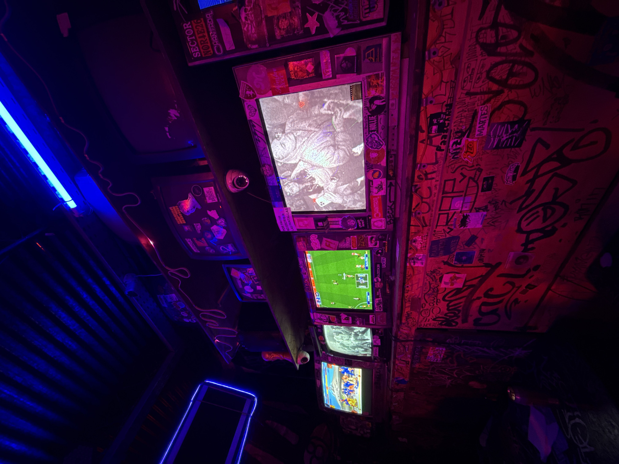 The image shows a view of a ceiling and walls covered in graffiti and stickers. Four monitors, encased with stickers and graffiti, are mounted on the ceiling, displaying different images: a black and white scene, a soccer game, a video game scene and a third video game. A blue neon light illuminates a corrugated ceiling. Text on the monitors' frames and adjacent surfaces are in Spanish, referencing locations like "CHANTADA" and "NOR-E26." A note, "POR FAVOR NO ES ESCRIBIR AS PANTALLAS" is partially visible on one monitor. The scene, possibly a bar or entertainment venue, is lit with blue and pink hues, suggesting a nightlife setting. The graffiti-laden decor and monitor content indicate an urban, possibly underground, culture. The setting appears to be within Buenos Aires, Argentina, given the Spanish text and general aesthetic associated with urban South American environments.