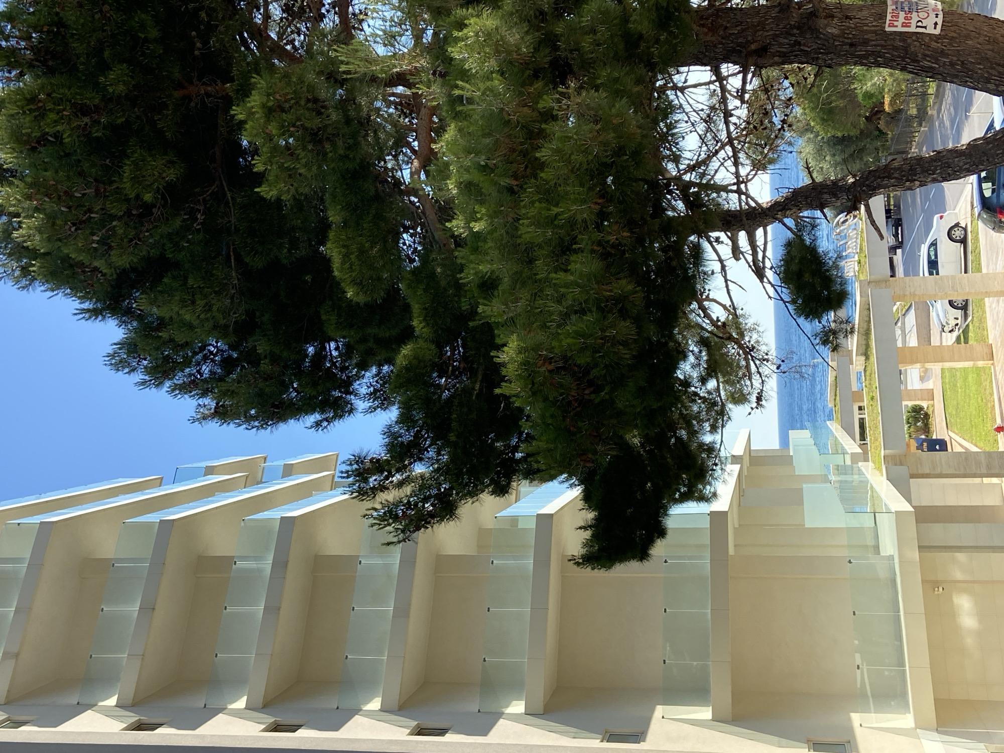 The image shows a building in Brežine, Montenegro. The architecture features white walls and glass panels. A large tree obscures part of the building and the sky. In the background, a road with cars and a glimpse of the sea are visible.