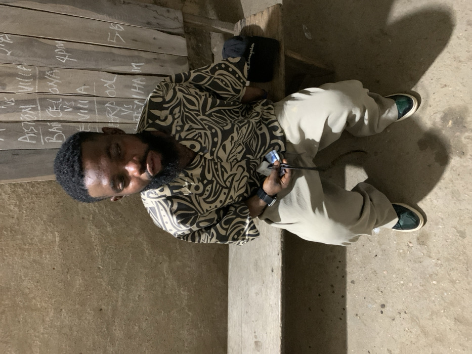 The image displays an adult male seated on a wooden bench in an establishment located in Okene, Nigeria. The individual has dark skin, a full beard, and short, dark hair. He is wearing a short-sleeved, collared shirt featuring a black and beige abstract pattern, light-colored trousers, and dark green shoes with white soles. A dark wristwatch is visible on his left wrist. In his left hand, he holds a small, light-colored object and a black cord. To his right, on the bench, rests a dark object with "Champion" written in white. The background on the left consists of a textured, light brown wall. To the right, a vertical wooden board, assembled from multiple planks, functions as a chalkboard. Handwritten in white chalk on this board are several football (soccer) match listings, including "ASTON VILLA VS", "BARCELONA VS", "SEVILLA", "LIVERPOOL VS", and "TOTTENHAM", along with times such as "4pm" and "56". The floor is a rough, light-colored concrete or packed earth surface. The scene is illuminated by an overhead light source, casting distinct shadows.