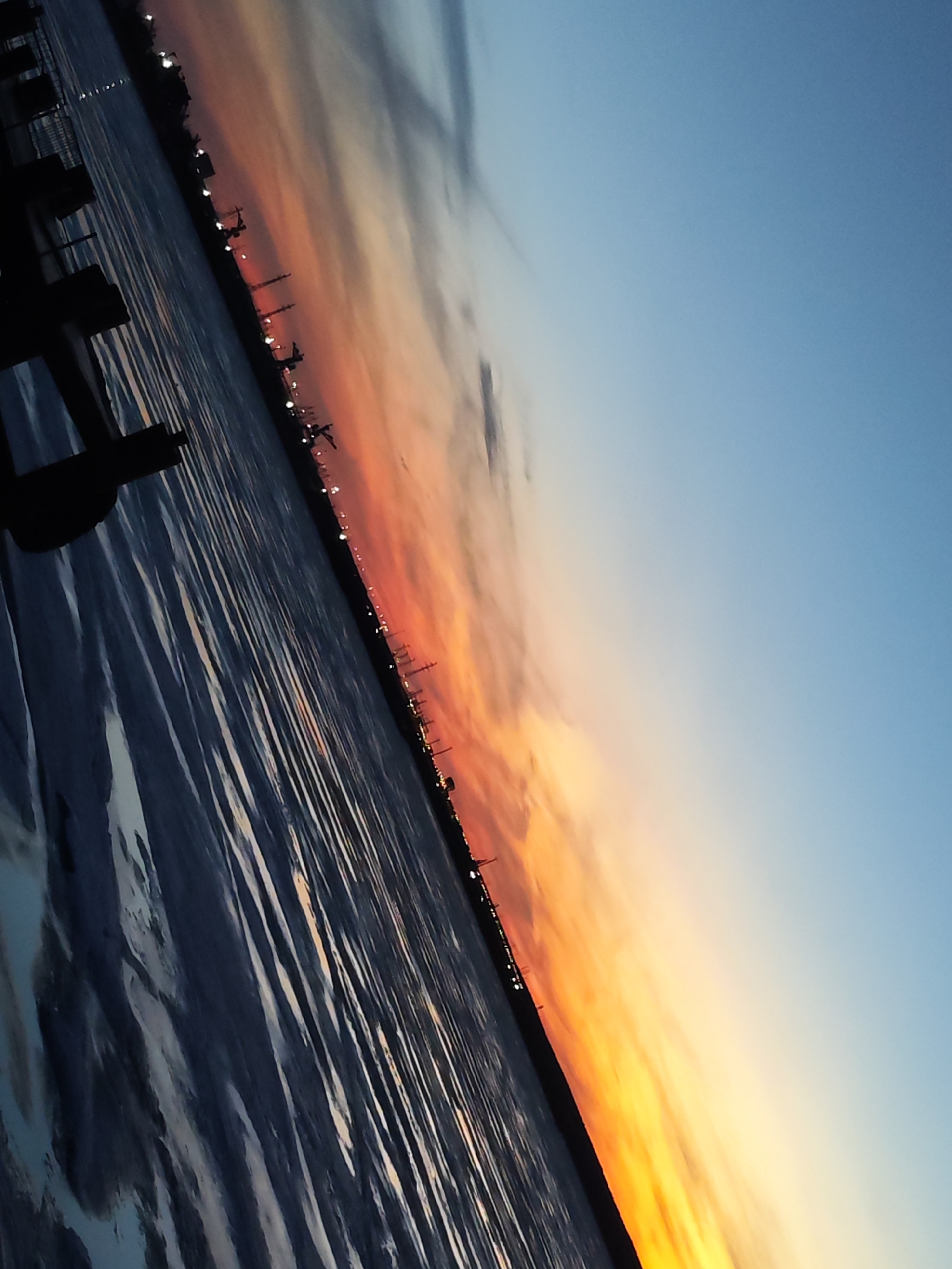 The image shows a sunset over a body of water in what appears to be the city of Chaikovsky, Russia. Silhouetted structures, likely industrial facilities, are visible along the horizon. The sky transitions from blue to orange and yellow hues near the horizon. The water's surface reflects the colors of the sky, creating linear patterns. A portion of a pier or dock is visible in the foreground.