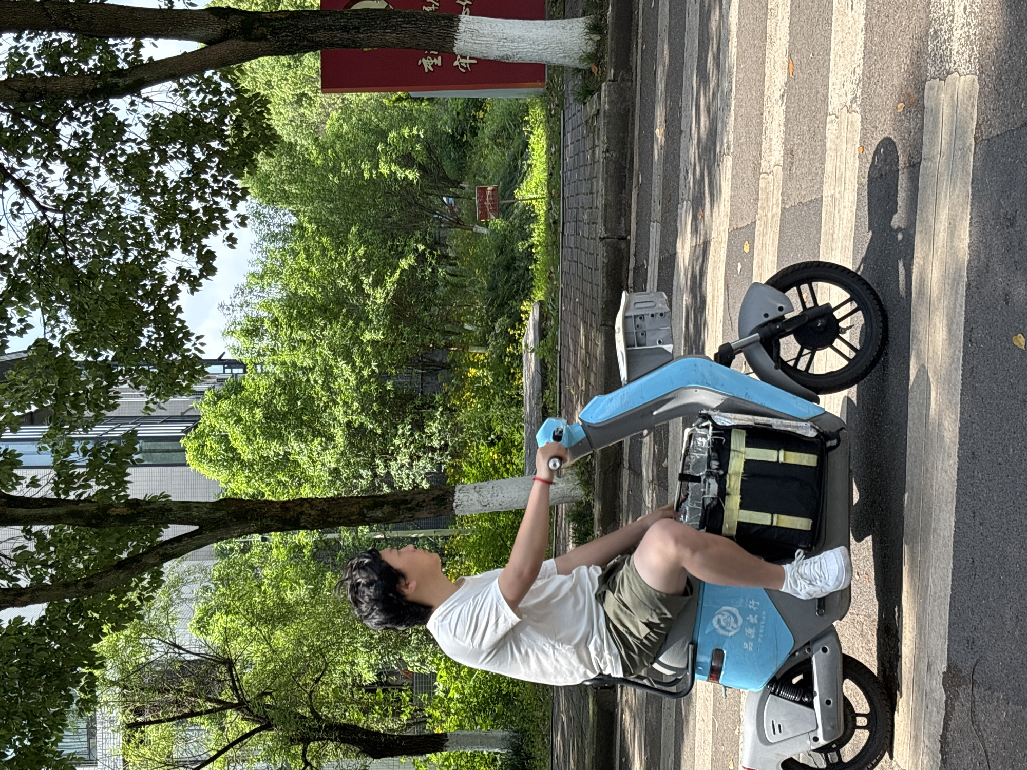 The image, set in Jiangjin, China, captures a person on a blue and gray electric scooter paused on a zebra crossing under bright daylight. The individual, appearing youthful with dark hair, wears a white t-shirt, olive green shorts, and white sneakers. Seated with a relaxed posture, they look slightly upwards or to the left, with their right hand resting on the scooter's handlebar.
The scooter features a black delivery bag secured to the rear. Distinct shadows cast by the person and the scooter across the asphalt road and white pedestrian stripes indicate strong sunshine.
The scene is surrounded by lush green foliage from numerous trees, some with trunks painted white at their bases. In the midground, behind the person and to the left, a large red signboard is partially visible, displaying discernible Chinese characters that read "福建" (Fújiàn). Further into the verdant background, another smaller, unreadable red sign can be glimpsed amidst the dense greenery. Building structures are faintly discernible through the trees on the left and right, suggesting an urban or semi-urban environment.
The electric scooter itself bears the Chinese characters "青桔单车" (Qīng Jú Dānchē), translating to "Qingju Bicycle" or "Qingju Shared Bike," and the English word "PENGUIN" on its side. The overall impression is a casual moment during a sunny day, possibly a brief stop at a crossing.