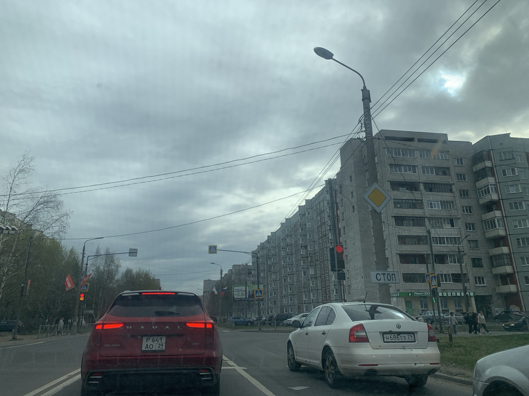 The image shows a street in Severodvinsk, Russia, on an overcast day. The view is from behind the wheel of a car, stopped in traffic. Ahead, a red Haval Jolion with the license plate "P 641 AO 29" is in front of the camera car. To the right of the Haval is a white Skoda Octavia with the license plate "H686 EB 29." Both cars are in the same lane, facing forward.
On the right side of the street, there is a tall apartment building with many windows. A street light and power lines run above the street. There is a diamond-shaped "yield" sign and a "STOP" sign in Russian ("СТОП") on the right side of the frame. Also visible is a crosswalk with people crossing the street. On the left side of the street, there are trees, street lights, and red flags. An advertisement of "-2,8" is visible on the left.
Several other cars are visible on the street. People are seen walking on the sidewalk on the right-hand side of the street.