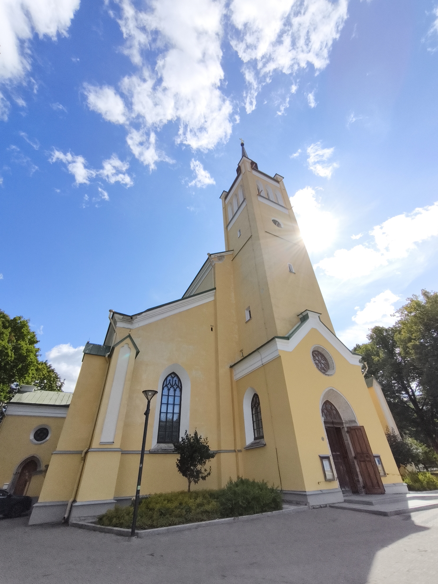 This is a low-angle, eye-level shot of the yellow exterior of the St. Charles Church (Kaarli kirik) in Tallinn, Estonia, on a bright, sunny day with blue skies and scattered white clouds. The church's tall bell tower dominates the right side of the frame, with the sun glinting brightly behind it. The main body of the church, painted a pale yellow with white trim, features gothic-style arched windows and a large, double-door entrance on the right. Green trees are visible behind the church on the right and left. A lamppost stands in front of the church to the left of the main entrance, next to a small bush. The ground in front is paved with grey asphalt.