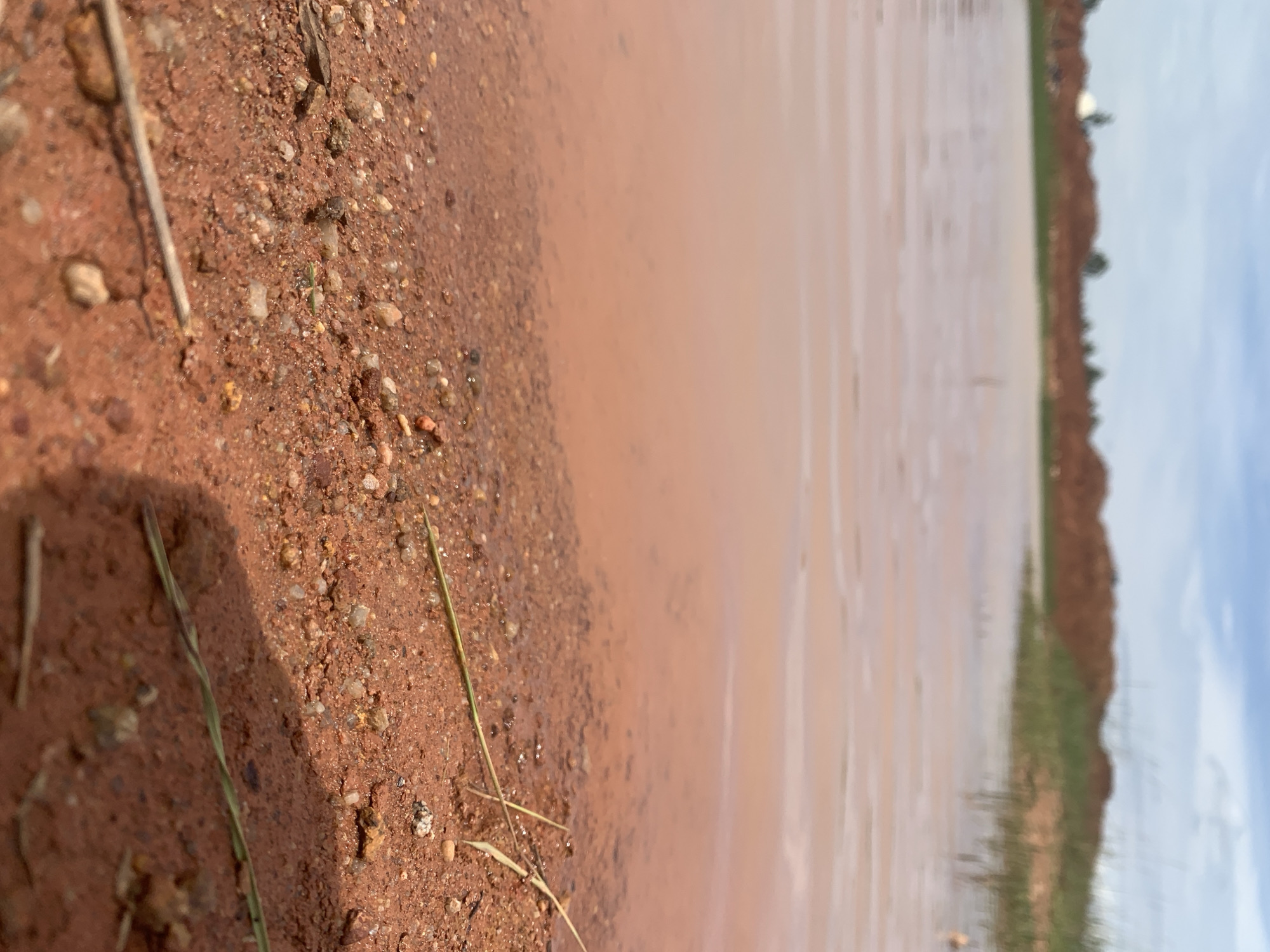 The image shows a shallow body of reddish-brown water, likely muddy, bordering a coarse sandy and gravelly shore. The water reflects the overcast sky, which is a mix of blue and white clouds. In the distance, there are blurred mounds of reddish earth, interspersed with green vegetation. The foreground is sharply focused on the textured shoreline, with small pebbles and dirt. A few dry grass stalks lie on the ground. The scene depicts a natural or disturbed landscape, possibly a construction site or a natural water accumulation area, in Rayfield, Nigeria. There are no people or animals visible in the image.