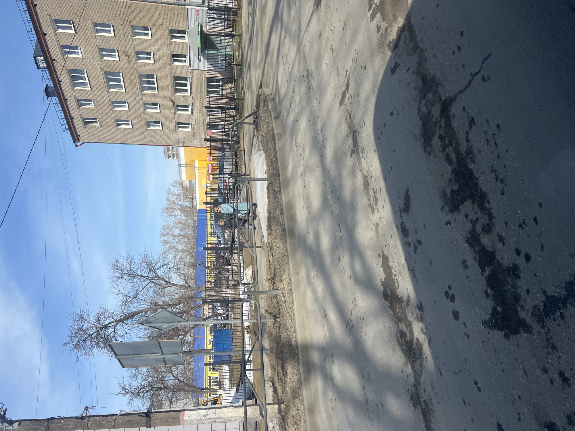 The image depicts a street scene in Chaikovsky, Russia. A multi-story brick building stands on the left. A fence runs along the sidewalk. Behind the fence, a blue and yellow structure is visible. Power lines stretch across the blue sky. The sidewalk and street show signs of wear and tear. A shadow is cast across the pavement. A road sign is present near the fence. A person is seen on a bicycle on the sidewalk. Trees with bare branches are visible.