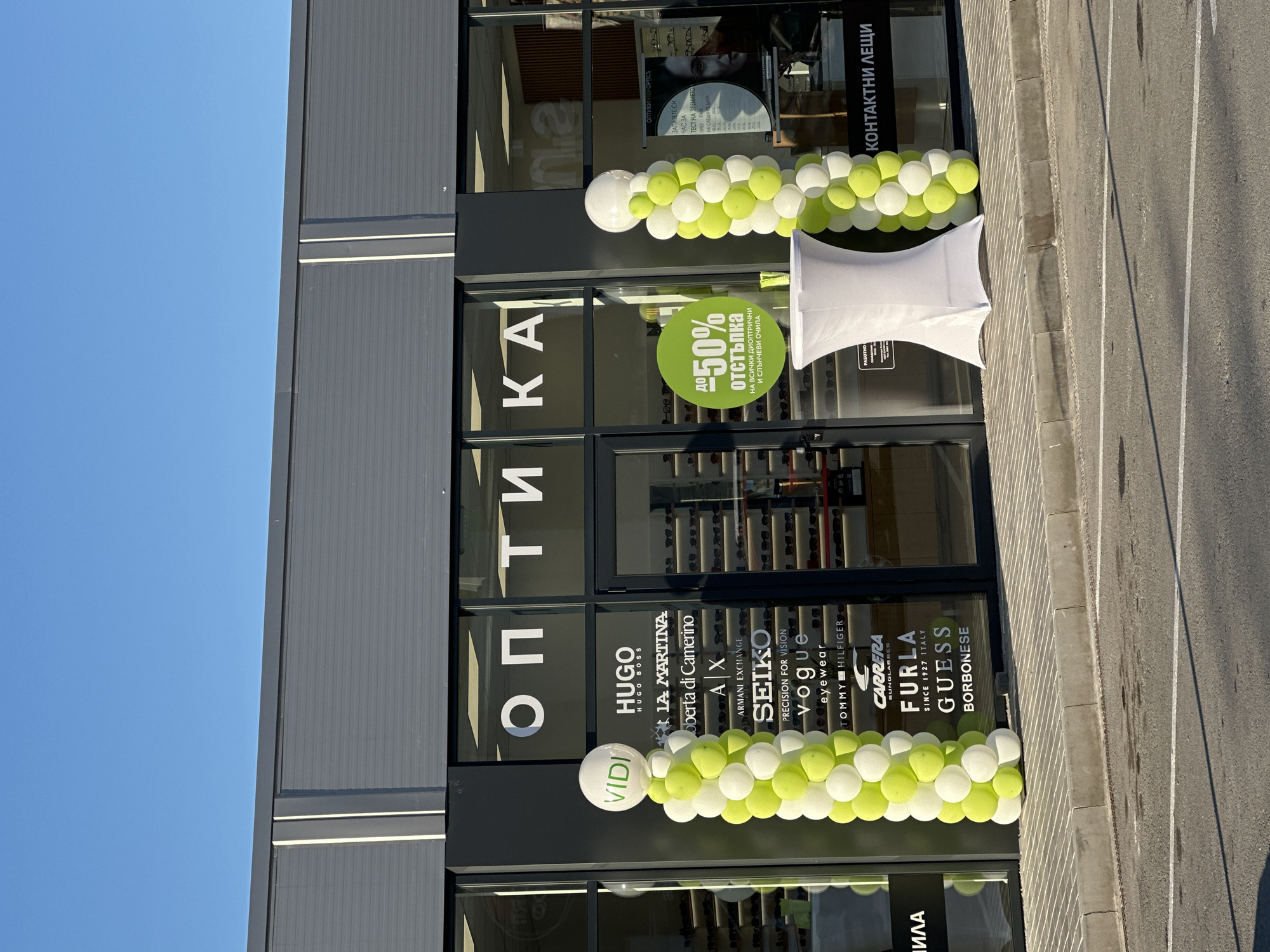 The image shows the exterior of an optics store called "ОПТИКА" in Veliko Tarnovo, Bulgaria. The storefront is modern with large glass windows displaying various eyewear brands like Hugo Boss, La Martina, Armani Exchange, Seiko, Vogue, Tommy Hilfiger, Carrera, Furla, Guess, and Borbonese. Two balloon arches, one on the left and one on the right, decorate the entrance. The left arch has a round sign at the top that says "VIDI" in green text. A green circular sticker on the window advertises "до 50% отстъпка" (up to 50% discount) on dioptric and sunglasses. A white draped table stands between the balloon arches. The environment is sunny with a clear blue sky. The store is located on a sidewalk next to a street with parking lines. The overall impression is of a grand opening or a special promotion event for the optics store.