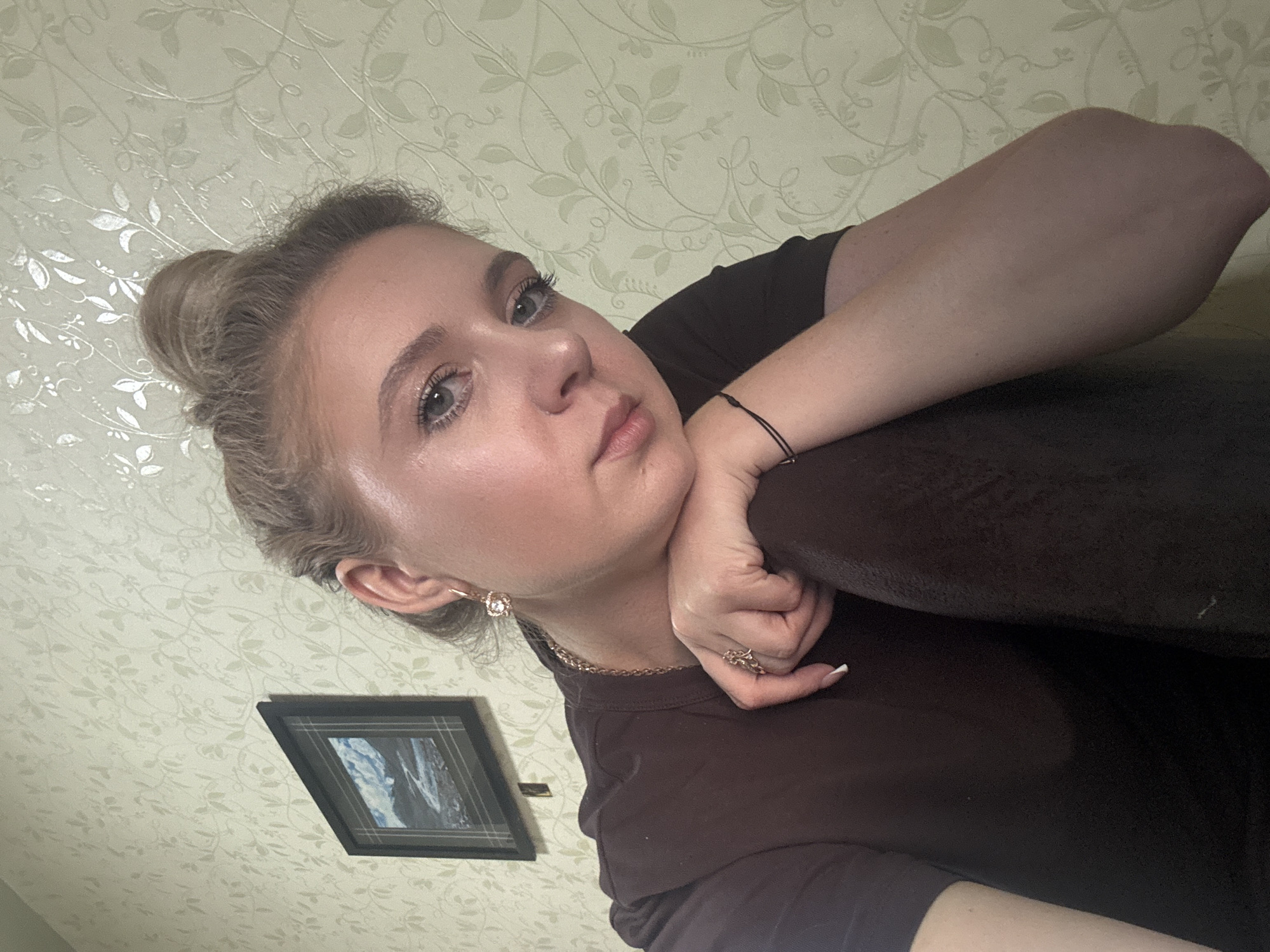 A young woman with blonde hair pulled up in a bun is leaning on her arm, looking forward. She is wearing a dark brown shirt and several pieces of jewelry, including earrings, a necklace, and rings. A framed picture of a landscape is hanging on the wall behind her. The wall has a light-colored, floral-patterned wallpaper. The image is a close-up shot, focusing on the woman's face and upper body.