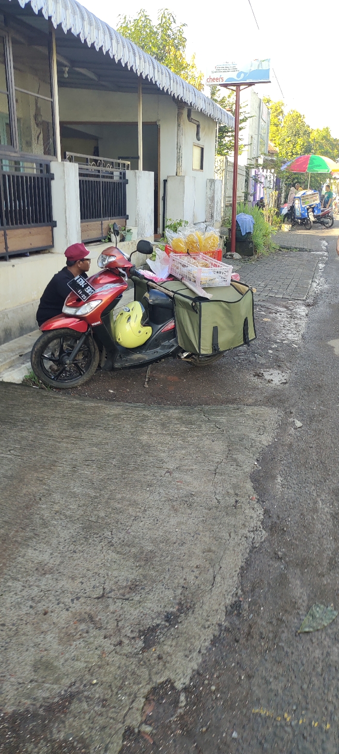 The image shows a man wearing a red hat and a black jacket sitting on a red motorcycle parked on a gray concrete sidewalk in front of a building. The motorcycle has a green bag strapped to the back and a yellow helmet is sitting on the seat. The man appears to be waiting for something. The building is a white concrete house with a corrugated metal roof. There is a sign that reads "cheers" on a pole in front of the building. There is another building further down the sidewalk with a corrugated metal roof. The sidewalk is cracked and dusty with some green vegetation in the background. The image appears to have been taken on a street in Kecamatan Lawang, Indonesia, at the coordinates (latitude: -7.847, longitude: 112.7). The image is likely taken in the early morning or late afternoon because of the angle of the sun.