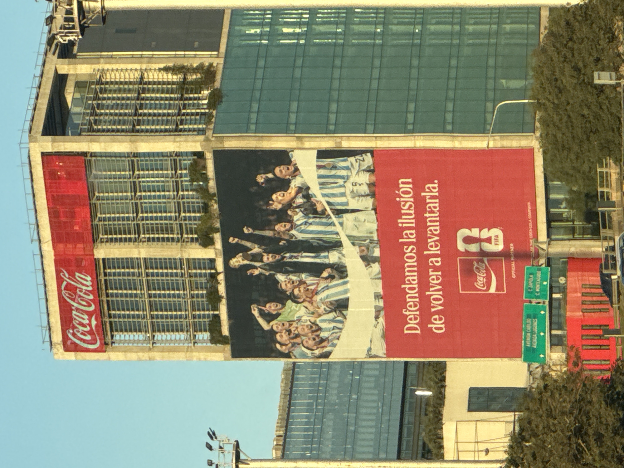 The image captures a large Coca-Cola advertising billboard affixed to the side of a tall building in Vicente López, Argentina. The advertisement features a graphic of the Argentinian national football team celebrating, with the text "Defendamos la ilusión de volver a levantarla" (Let's defend the illusion of rising again). The Coca-Cola logo is prominently displayed, along with the FIFA logo. The background shows a mix of modern architecture and greenery, typical of an urban environment. Street signs are visible indicating nearby locations. The sky is clear, suggesting daytime.