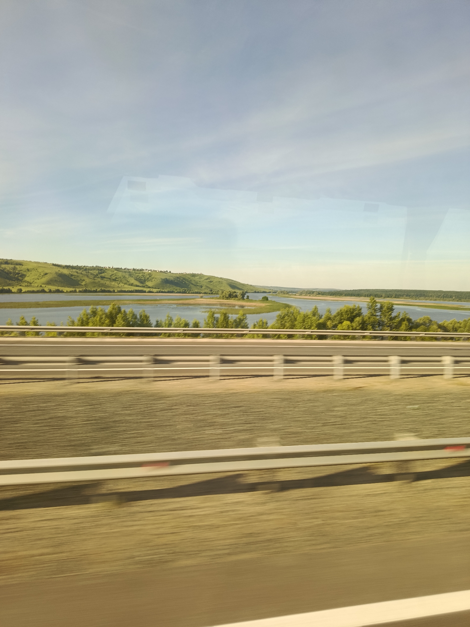 The image captures a scenic view of a wide river with lush green hills in the background. A tree-lined embankment separates the river from a road, which features a white guardrail. The sky is a clear, pale blue with some wispy clouds. The overall impression is one of a tranquil, natural landscape, possibly captured from a moving vehicle due to the slight motion blur in the foreground. The location is identified as Nizhniye Vyazovye, Russia.