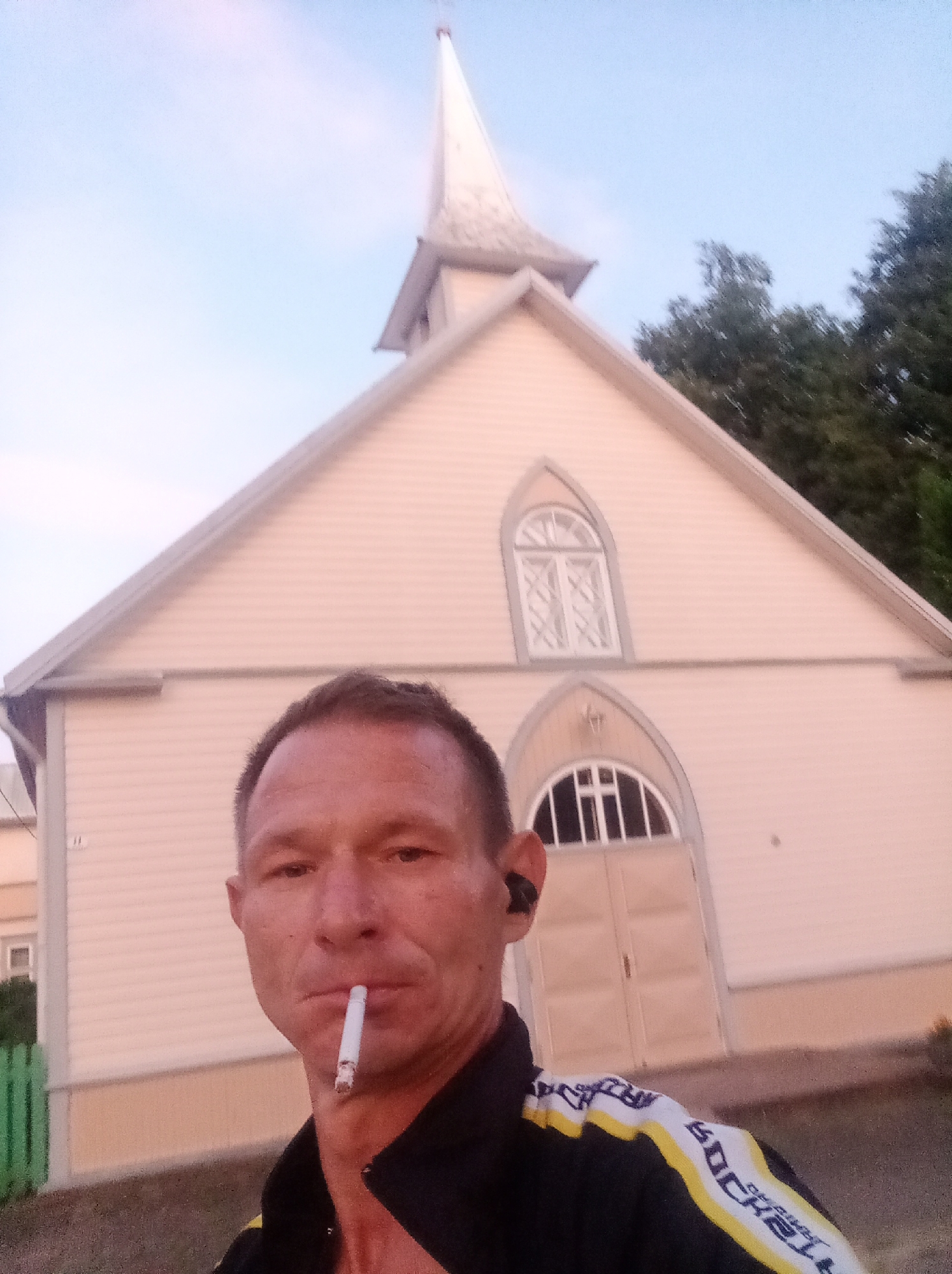 A man stands in front of a light pink, wooden church with a white steeple. The man has short, brown hair and is looking at the camera with a cigarette in his mouth. He is wearing a black jacket with yellow and white stripes. The church has arched windows and doors, and a light blue sky is visible in the background. In the foreground, there is a green fence. The location is believed to be the Tapa Evangelical Lutheran Church in Tapa, Estonia.