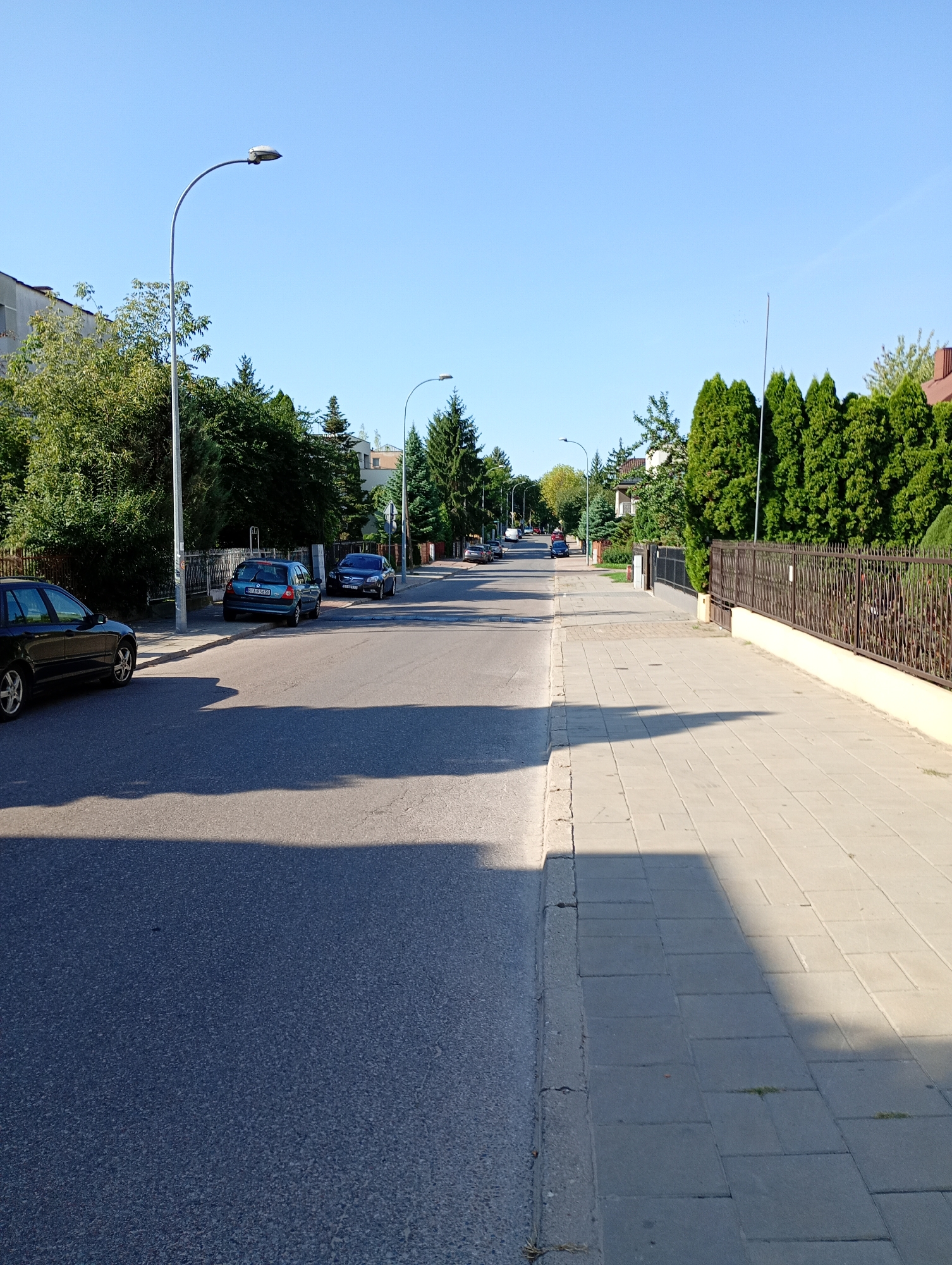 This is a daytime, eye-level, full shot of a residential street in Białystok, Poland. The street is paved with asphalt, and there is a concrete sidewalk on the right side of the image. Several cars are parked on the side of the street, and there are trees and houses lining the road. The sky is clear and blue. A street lamp is visible on the left side of the image. The shadows indicate that the sun is low in the sky, suggesting it is either early morning or late afternoon.