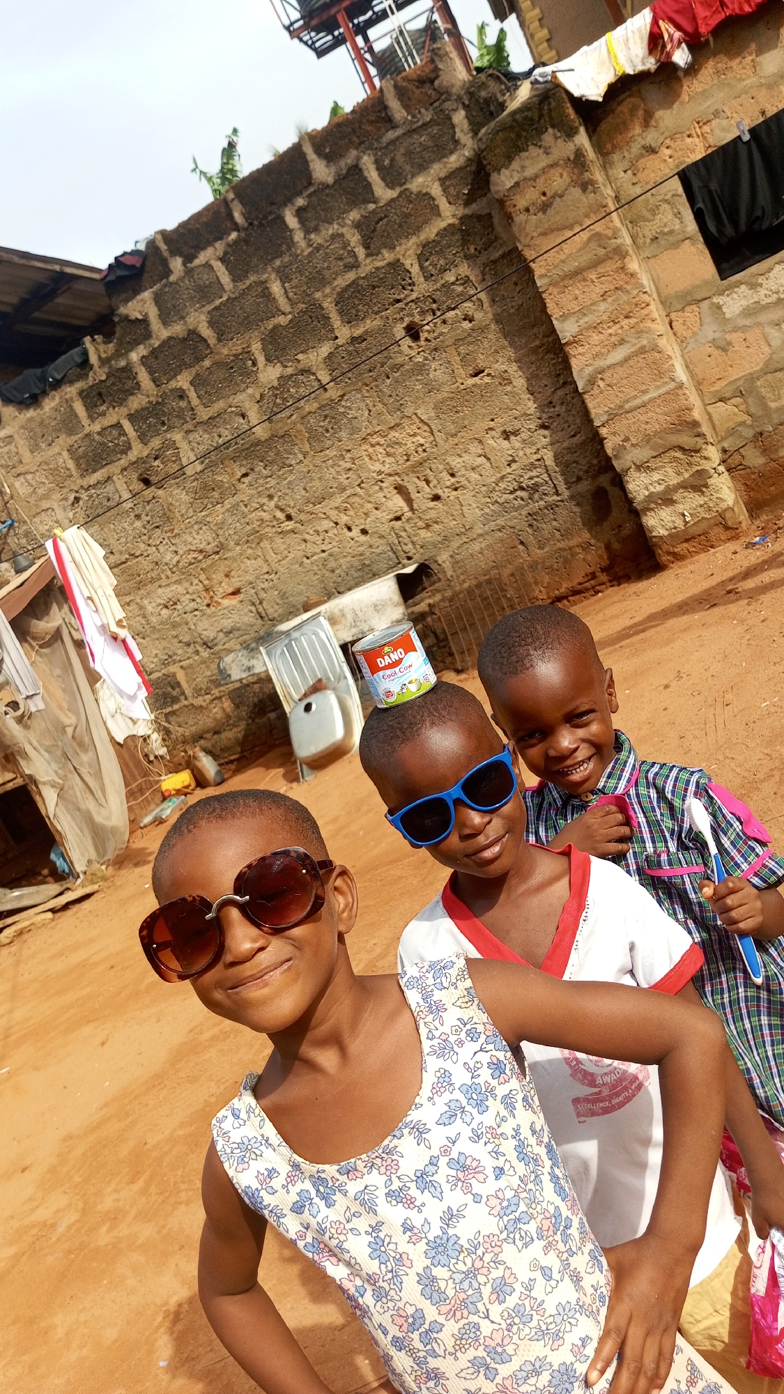 In Nkpor, Nigeria, three young children pose outdoors. The child in the foreground wears a floral-patterned sleeveless dress and oversized sunglasses. The child behind her wears a white t-shirt with red trim and blue sunglasses, balancing a can of "DANO Cool Cow" milk powder on his head. The third child, in the background, wears a plaid shirt and smiles, holding a blue toothbrush. Behind them, a block wall is visible, along with clotheslines, a metal sink, and other household items. The ground is reddish-brown dirt.