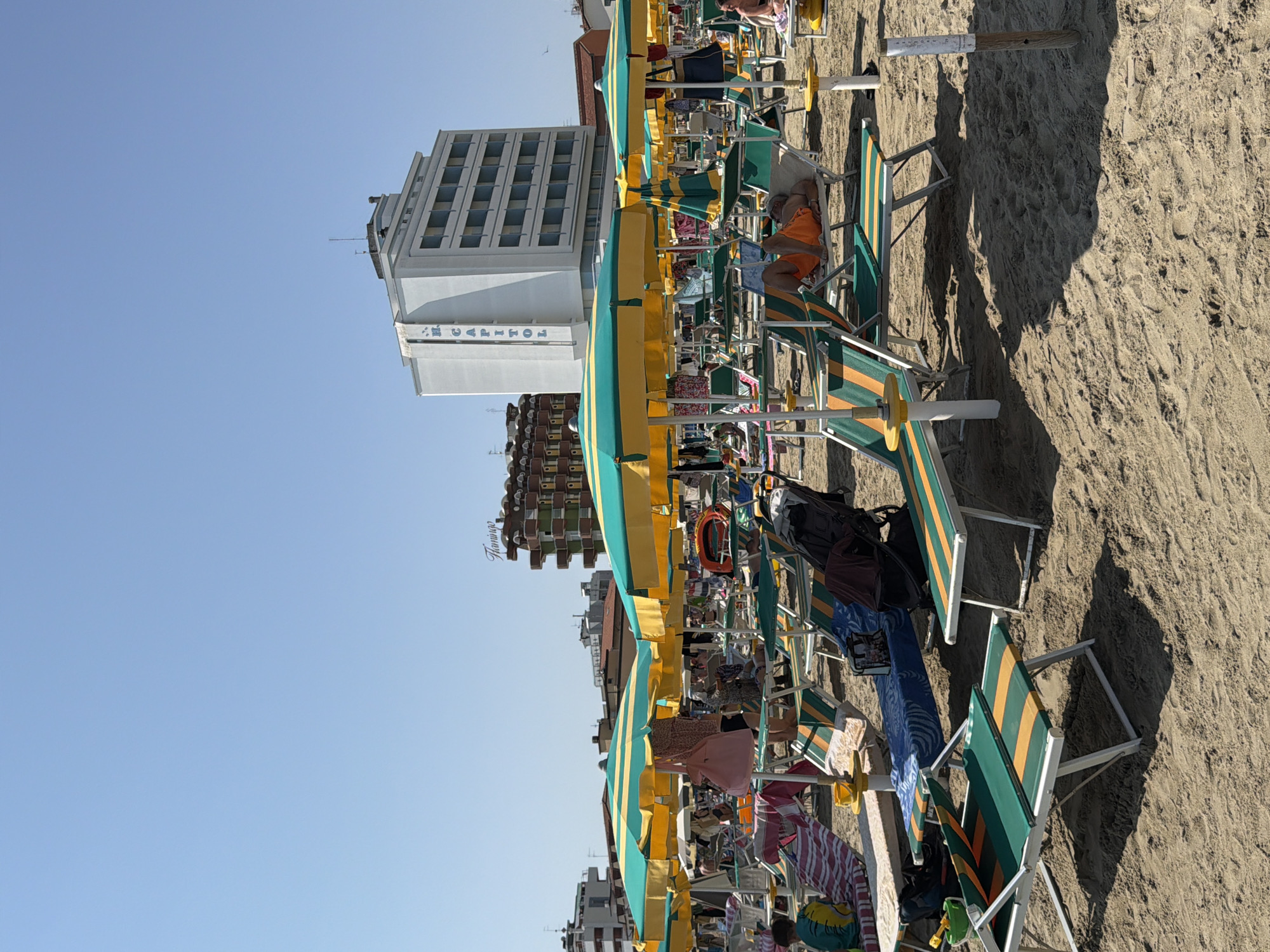 The image depicts a crowded beach scene in Gatteo, Italy, under a clear blue sky. The foreground and midground are dominated by rows of empty and occupied beach chairs and umbrellas, characteristic of a summer day at the seaside. The umbrellas are predominantly green and yellow striped, with some solid green ones visible. Various beach towels and personal items are spread across the chairs. Several people are visible, some relaxing on loungers, others walking or standing under umbrellas. In the background, there are multi-story buildings, including a prominent white building with "H. CAPITOL" written vertically on its facade, suggesting the presence of hotels or other accommodations. Another building with a sign that appears to read "Flamingo" is also visible. The beach itself is sandy, with shadows cast by the umbrellas and chairs indicating a bright, sunny day. The overall impression is one of a bustling tourist destination.