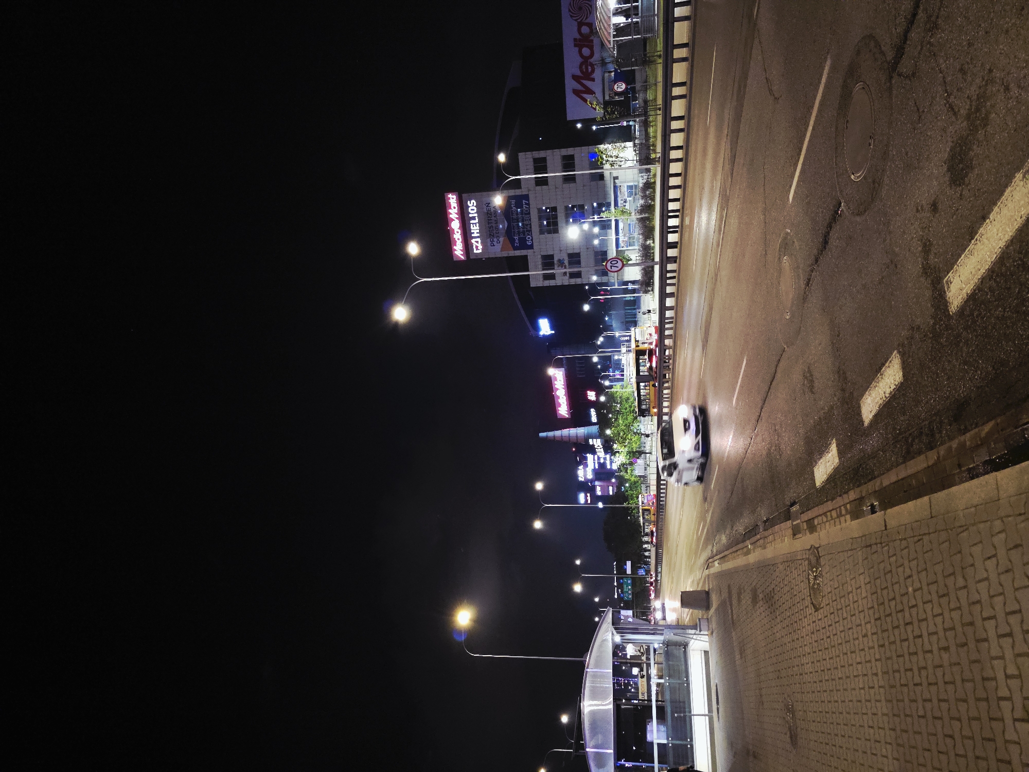 The image captures a nighttime scene in Warsaw, Poland, dominated by a street view illuminated by streetlights and commercial signage. The street itself is dark and wet, with visible lane markings and some surface imperfections. A white car is in motion on the road.
Notable signage indicates "Media Markt" and "Helios," suggesting the presence of retail outlets or entertainment venues. A bus stop is visible in the foreground. The sidewalk is constructed of interlocking pavers. The sky above is dark, typical of a night scene.