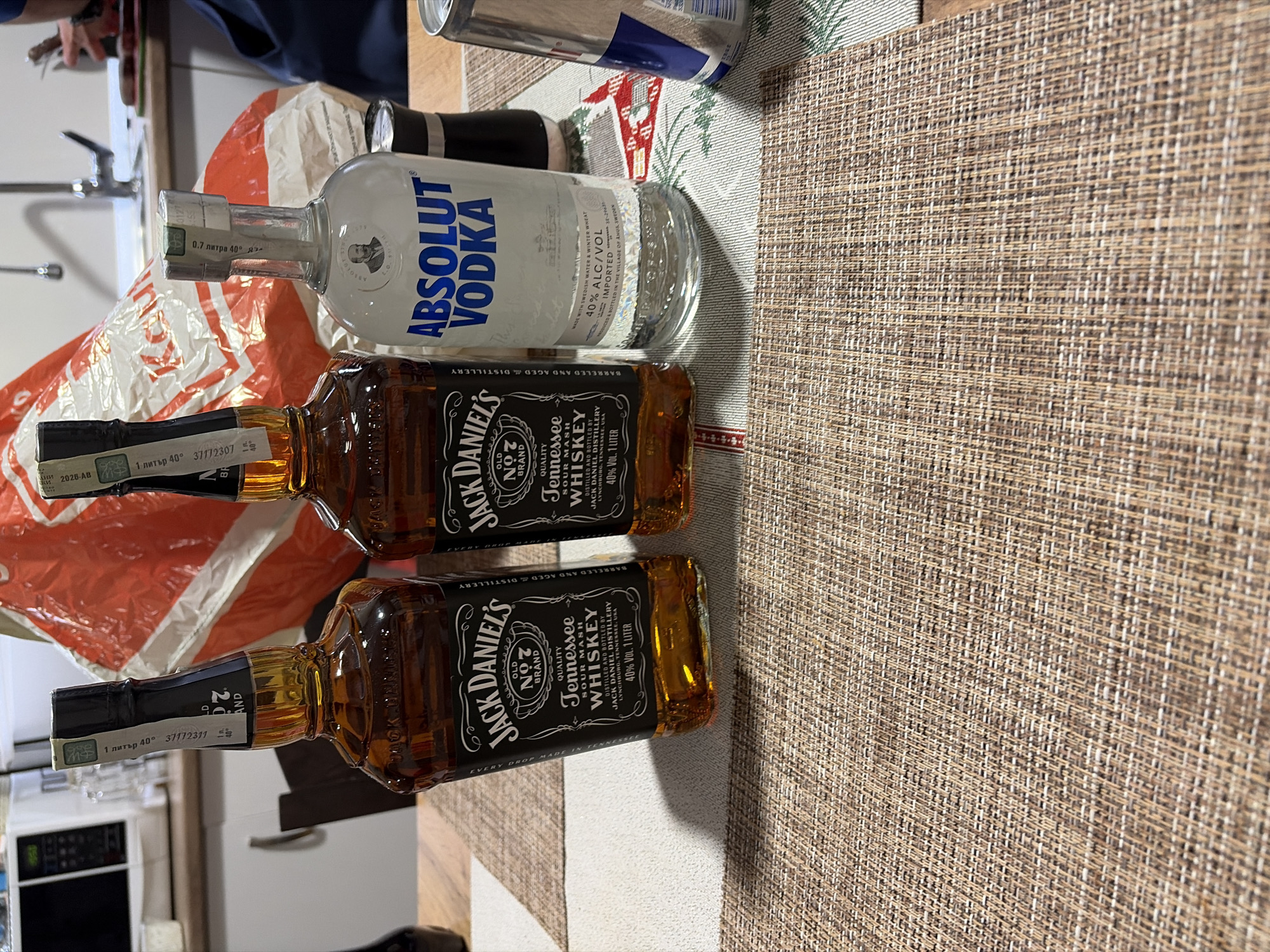 The image shows two bottles of Jack Daniel's Tennessee Whiskey (1 liter, 40% ABV) and one bottle of Absolut Vodka (0.7 liters, 40% ABV) resting on a woven placemat. A can of Red Bull is visible behind the vodka bottle. A red and white plastic bag is partially visible behind the bottles. The setting appears to be a kitchen counter, with a microwave visible in the background. The bottles are likely located in a kitchen in an unknown location. There is no indication of location of Madzherito, Bulgaria.