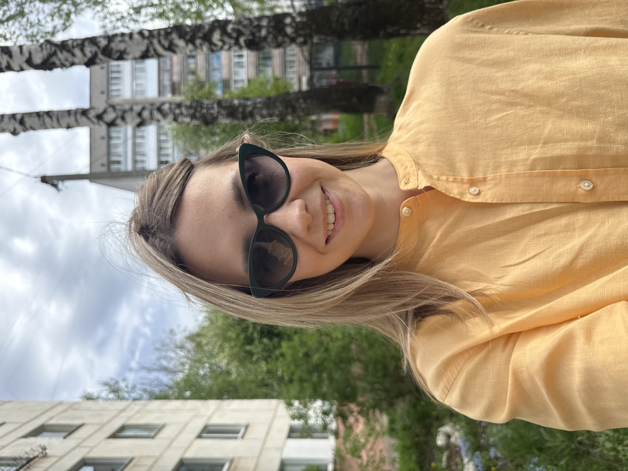 A young woman with blonde hair is posing for a photo outdoors. She's wearing sunglasses and an orange collared shirt. The background shows a residential building and trees, characteristic of a city setting. Given the request to locate it in Chaikovsky, Russia, one can assume this photograph was taken there, in a public space like a park or residential area.