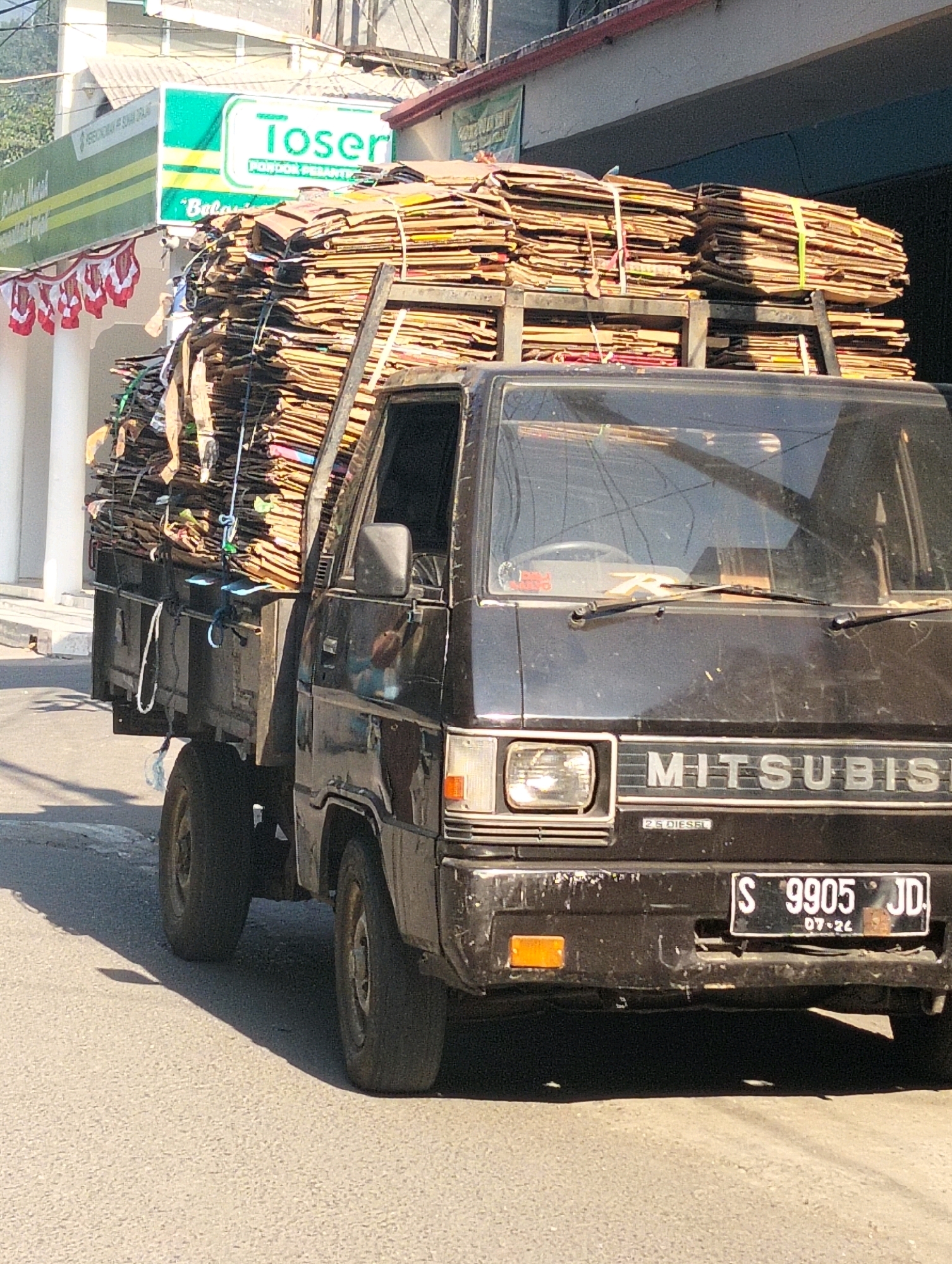 In Sambipondok, Indonesia, a black Mitsubishi truck is parked on a street. The truck is heavily loaded with stacks of flattened cardboard boxes, secured with ropes. The vehicle has a license plate reading "S 9905 JD" and "07-24." In the background, a building with a green and white sign that says "Toser" is visible, along with red and white decorative banners hanging from a railing. The street is paved with asphalt, and sunlight casts shadows from the truck and an unseen object onto the road. The scene appears to be a commercial area of the city.