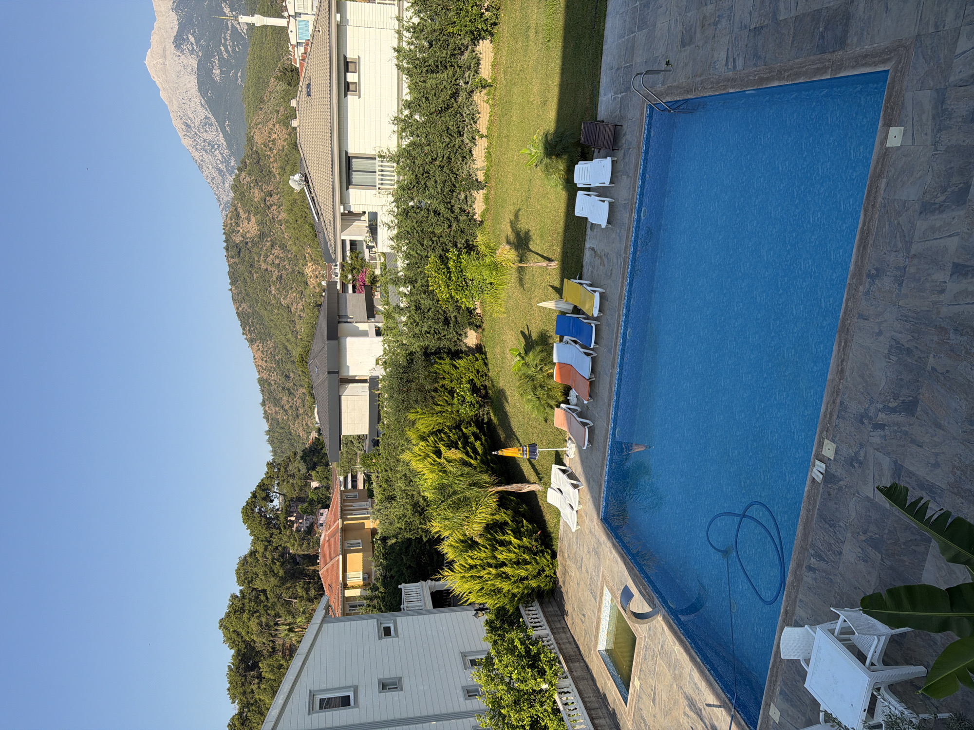 This is a high-angle, vertical, exterior shot of a swimming pool area at a residential property, likely a villa or a resort. The pool is a vibrant blue and appears to be well-maintained, with a hose for cleaning visible coiled in the water. Several lounge chairs are arranged around the pool, some occupied by what appear to be people or dolls, suggesting a leisurely day. The surrounding area is lush and green, with manicured lawns, trees, and shrubs. In the background, there are several buildings, including houses and a structure with a minaret, hinting at a Mediterranean or Middle Eastern location. Towering mountains dominate the skyline, creating a dramatic backdrop. The sky is clear and blue, indicating fair weather, and the lighting suggests it is daytime, likely mid-morning or afternoon, given the shadows. The scene evokes a sense of relaxation and vacation.
