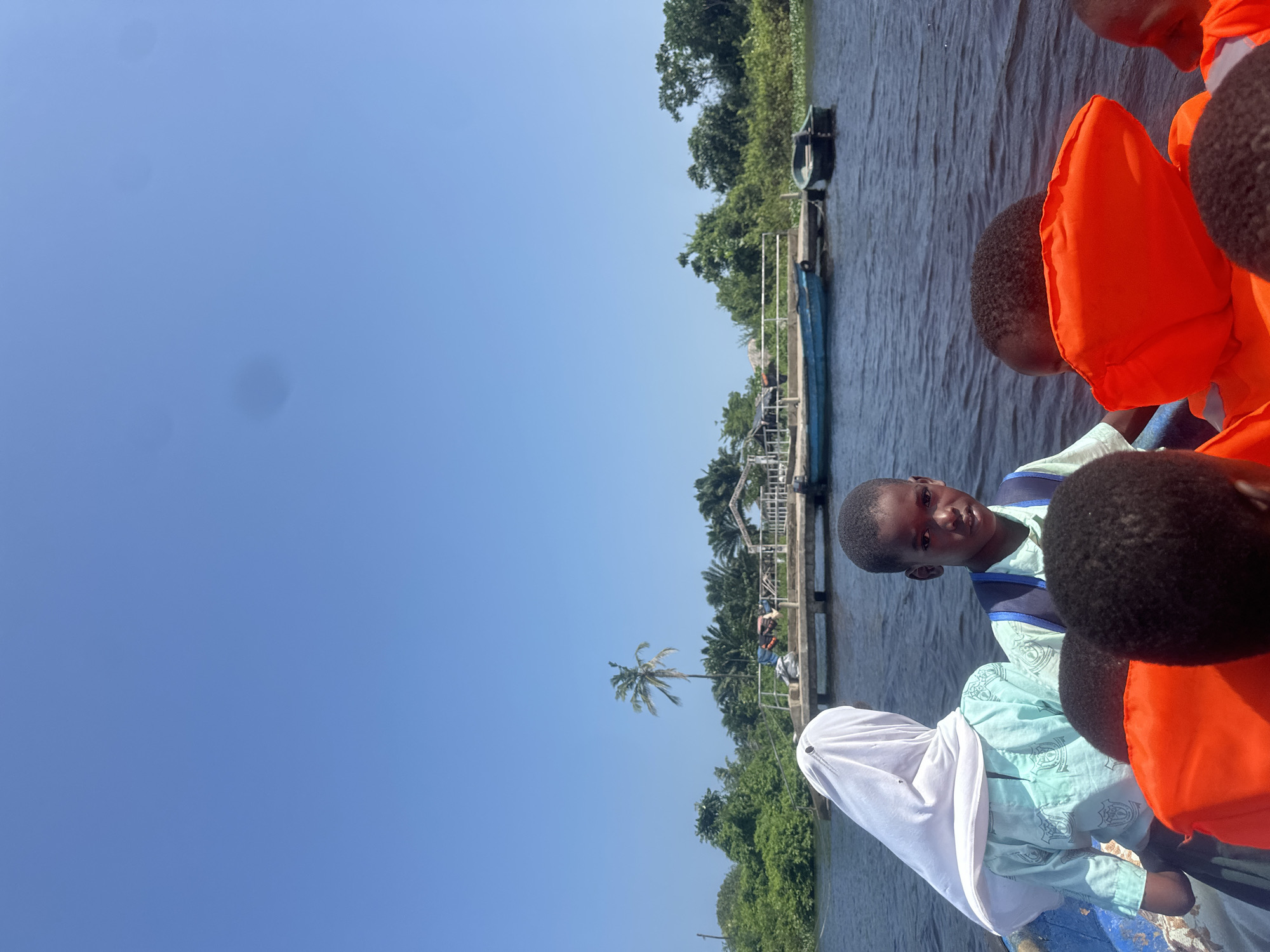 In Badagry, Nigeria, a group of children are on a boat trip. The boat is in a body of water, and a wooden dock with a few structures is visible in the background. Lush green vegetation lines the shore, with palm trees and other tropical plants. The sky is clear and blue. Some of the children are wearing bright orange life vests. One boy in the foreground is looking directly at the camera. Another child is wearing a light blue shirt and a white headscarf. The overall scene suggests a day of travel or excursion on the water.