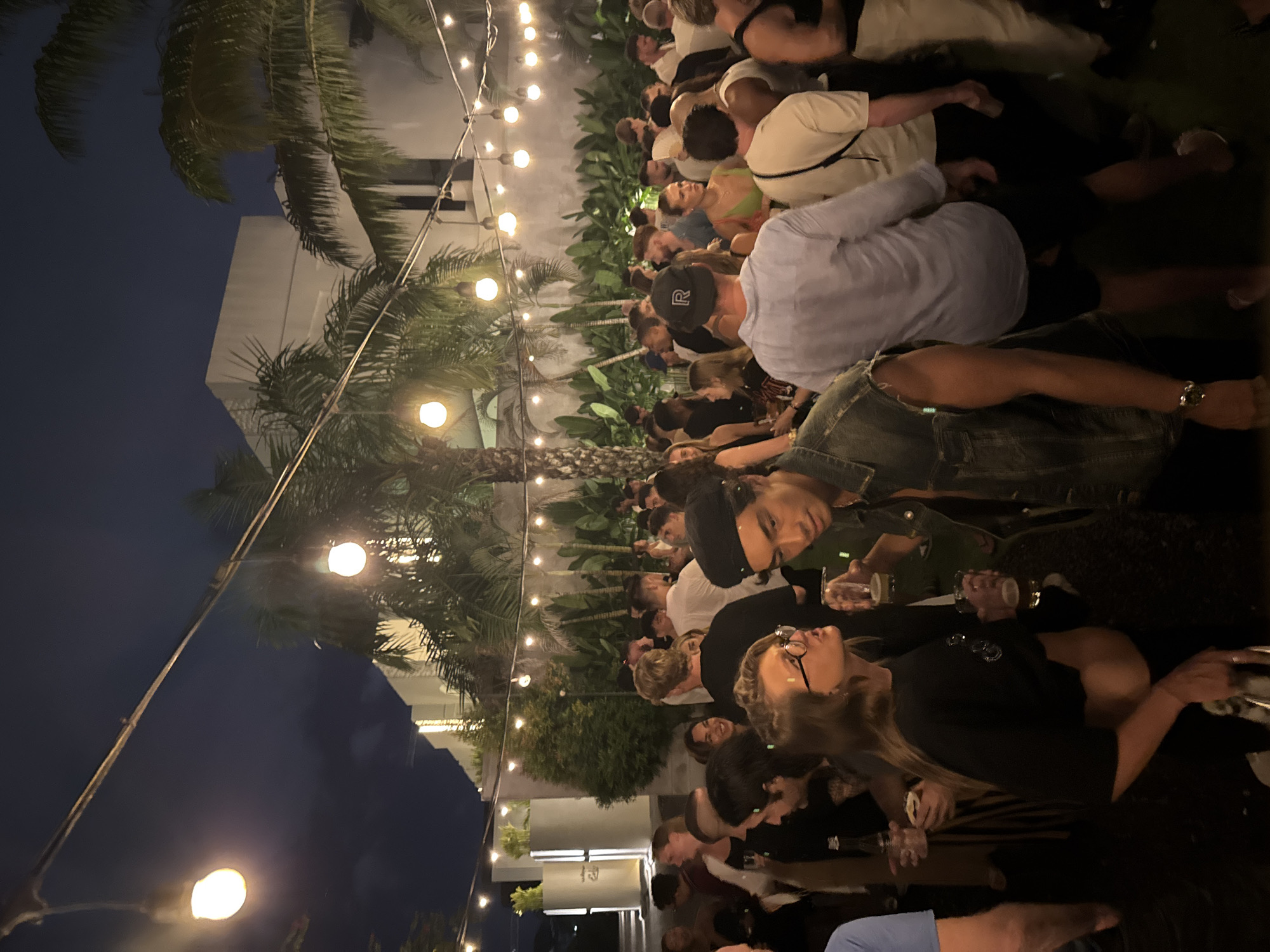 This media file captures a lively outdoor night scene at what appears to be a bar, restaurant, or social gathering in North Kuta, Indonesia. The image is taken at night, with a dark sky visible, and the area is brightly illuminated by numerous string lights strung overhead, creating a festive and warm ambiance.
The scene is crowded with many people, primarily young to middle-aged adults, standing and mingling. They are dressed in casual attire, including t-shirts, light shirts, and denim. Many individuals are holding drinks in glasses or cups, suggesting a social event focused on refreshments and conversation. People are seen facing each other, engaged in discussions and enjoying the evening. Emotions appear generally positive, with relaxed postures and smiles visible on some faces.
Notable subjects include several palm trees and lush green foliage lining the back of the gathering area, reinforcing the tropical location. A light-colored building with multiple stories is visible in the background. In the foreground, a man wears a black baseball cap with a white "R" logo. The string lights are a dominant feature, casting a warm glow and defining the atmosphere of the outdoor venue. The overall impression is that of a bustling and enjoyable evening event in a tropical setting.