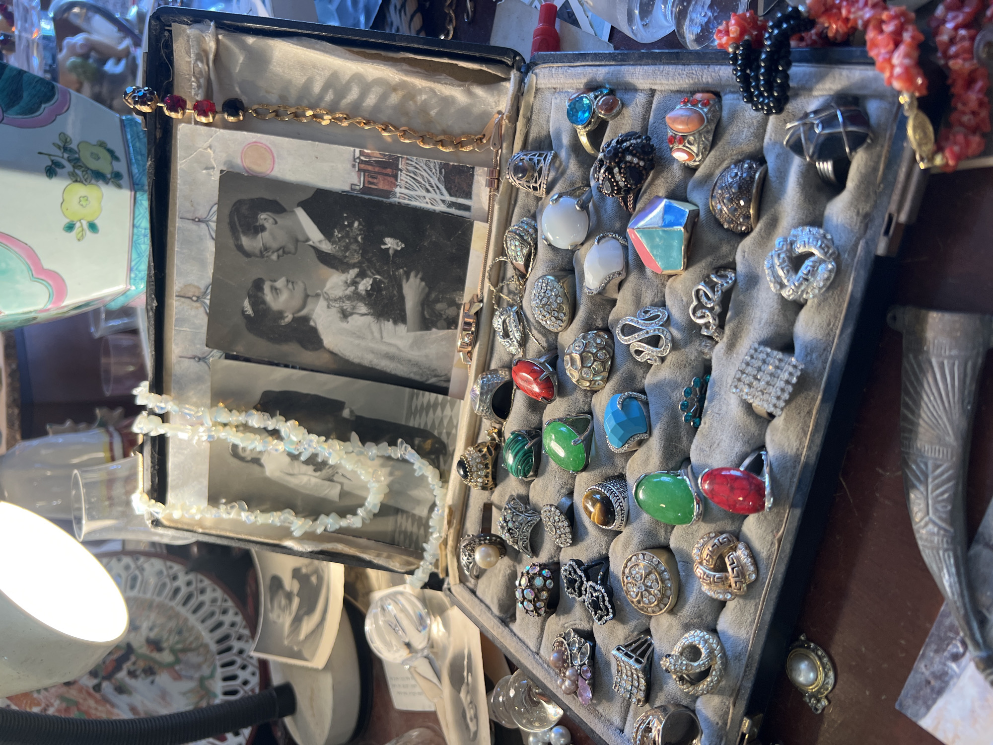 The image shows an open jewelry box filled with an assortment of rings, bracelets, and necklaces. The box is divided into two sections. The left section contains photographs, a pearl necklace, and a gold chain bracelet with colorful stones. The right section is filled with an array of rings with varying designs, colors, and stones, displayed on a grey velvet surface. In the background, miscellaneous objects are visible, including glass items, jewelry pieces, and parts of other decorative objects. The location and the city are unknown.