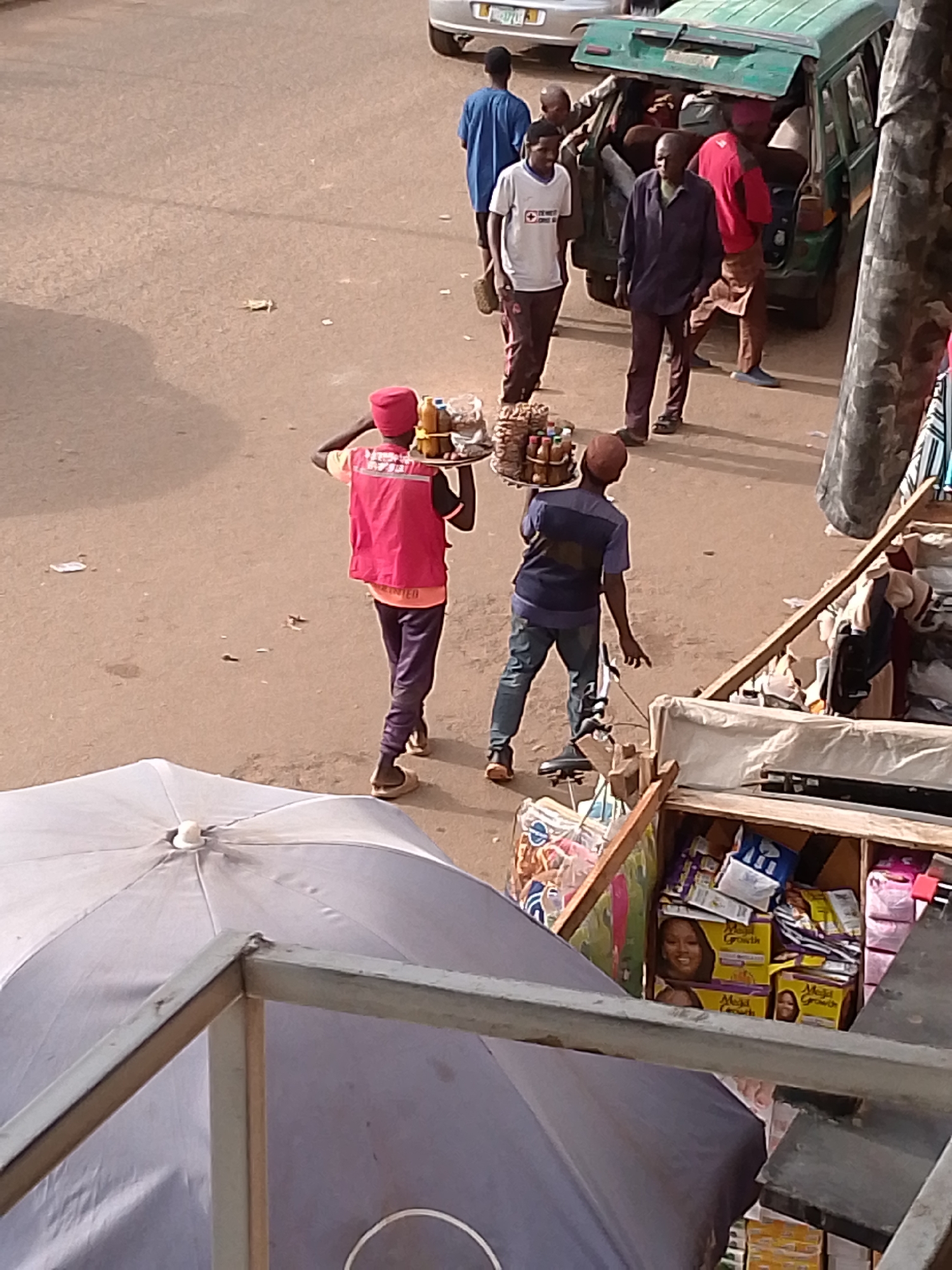 A high-angle, eye-level medium shot captures a street scene in an urban environment. In the foreground, a gray umbrella is partially visible, along with a metal railing. To the right of the umbrella, a wooden stall is filled with packaged goods, including boxes featuring images of women and the text "Mega Growth." Two individuals are the primary subjects. The first, wearing a red vest and a red cap, is carrying a tray laden with bottles and snacks. The second individual, wearing a blue t-shirt and jeans, is walking away from the camera. In the background, a green vehicle with its rear door open is parked, and several other people are milling around it. The ground is paved, and litter is scattered across the surface.