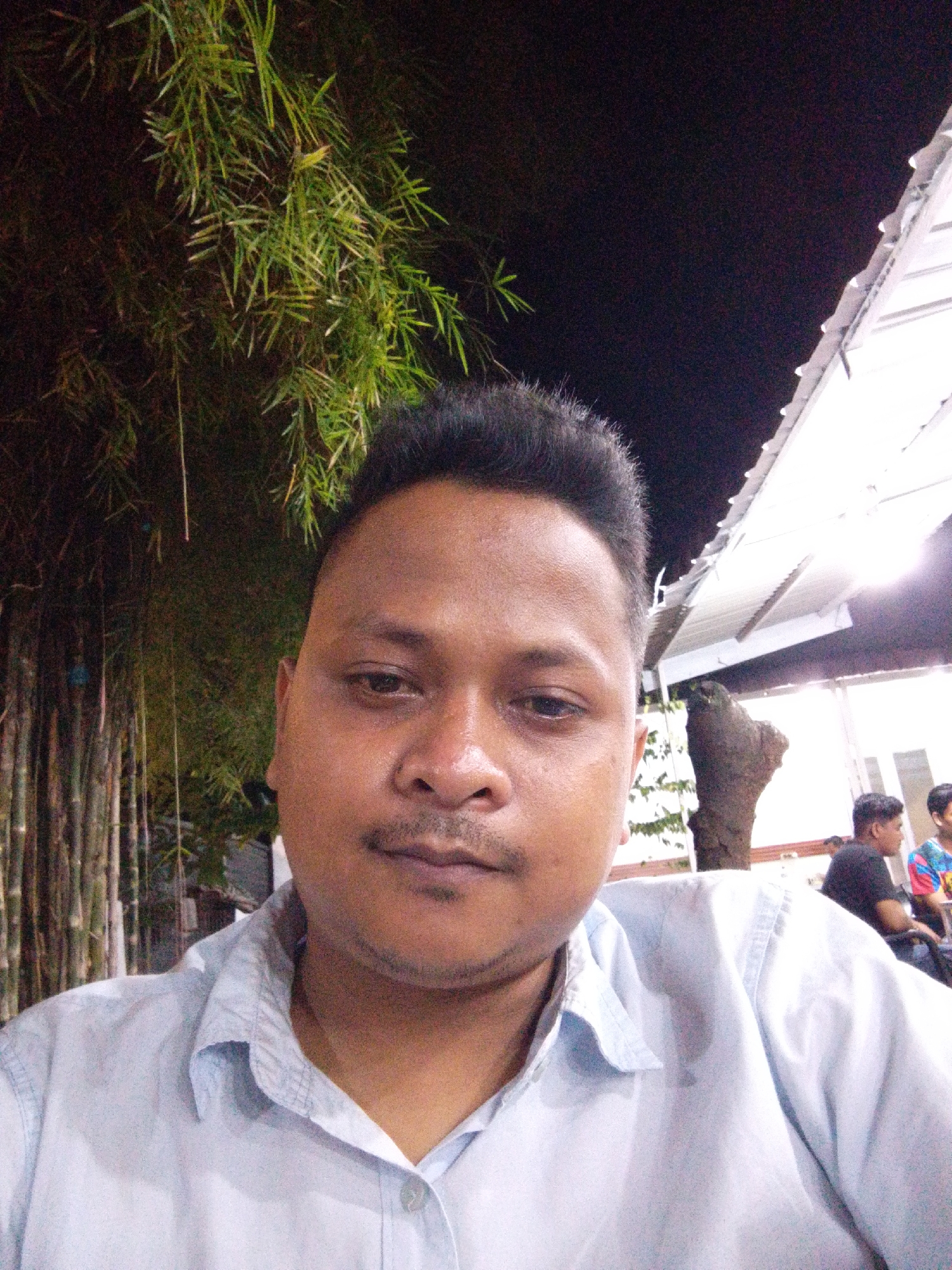 The image shows a medium shot of a man with short black hair and a mustache, wearing a light blue button-down shirt. He appears to be taking a selfie outdoors at night.
Behind him on the left is dense green foliage, possibly bamboo. To the right, there's a partially visible structure with a white roof, suggesting a building or shelter. Further in the background, other people are seated.
The lighting suggests it is artificial, likely from nearby buildings or structures. The location appears to be a public space or outdoor dining area in Lhokseumawe, Indonesia, although specific landmarks are not clearly visible.