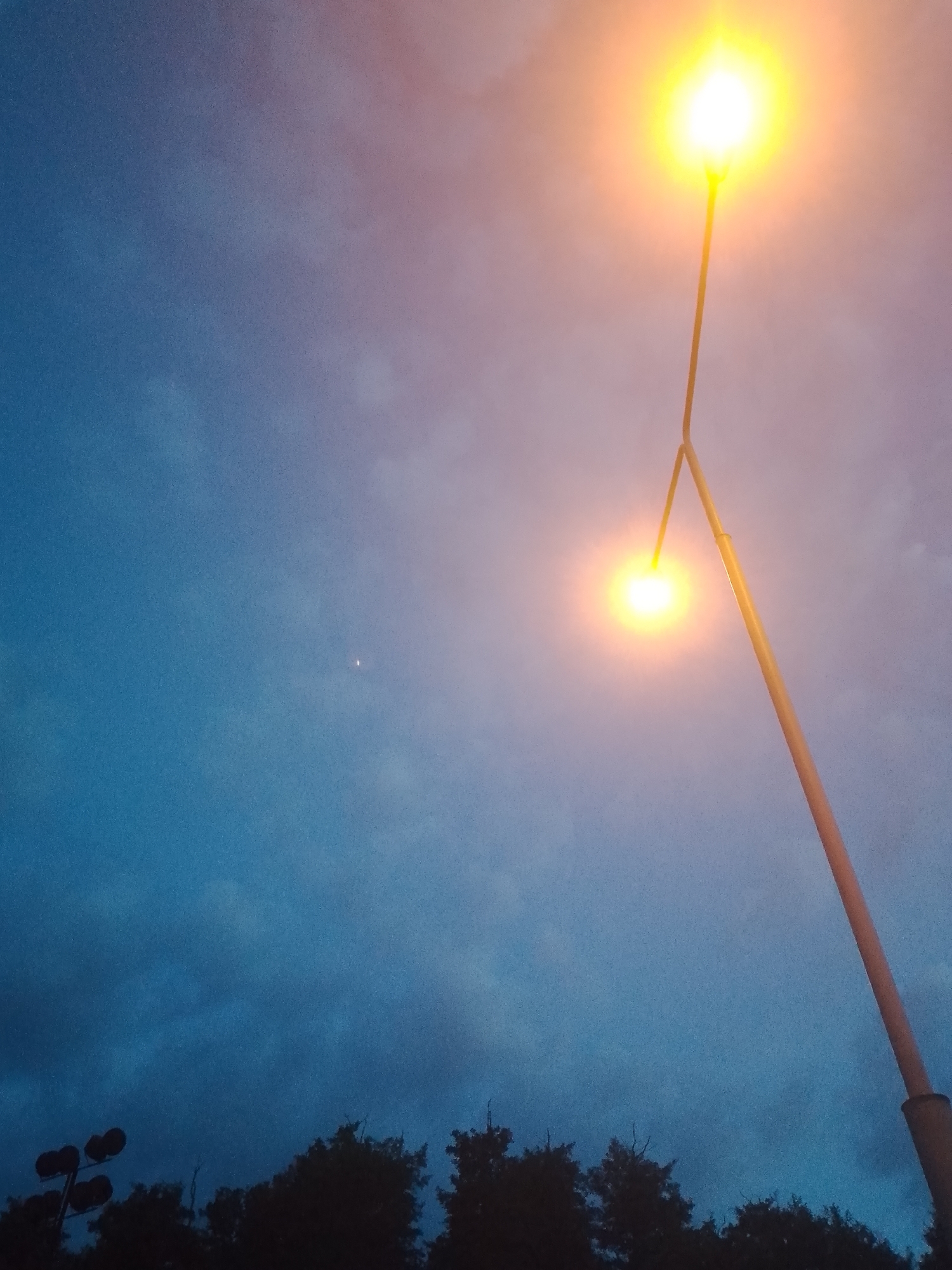 This is a photograph taken at dusk, capturing a sky with hues of blue, pink, and orange. The image features a tall street light with two bright yellow lamps, positioned on the right side of the frame. The lower lamp is closer and more prominent, while the upper lamp is slightly higher and more diffused. In the distance, a small, bright white speck is visible in the sky, potentially a star or a distant aircraft. The bottom of the image shows the dark silhouettes of trees and a cluster of stadium lights on the left. The overall environment suggests an outdoor setting in a city, possibly a park or a sports facility. The time of day and the presence of street lights indicate evening. There are no people visible in the image.