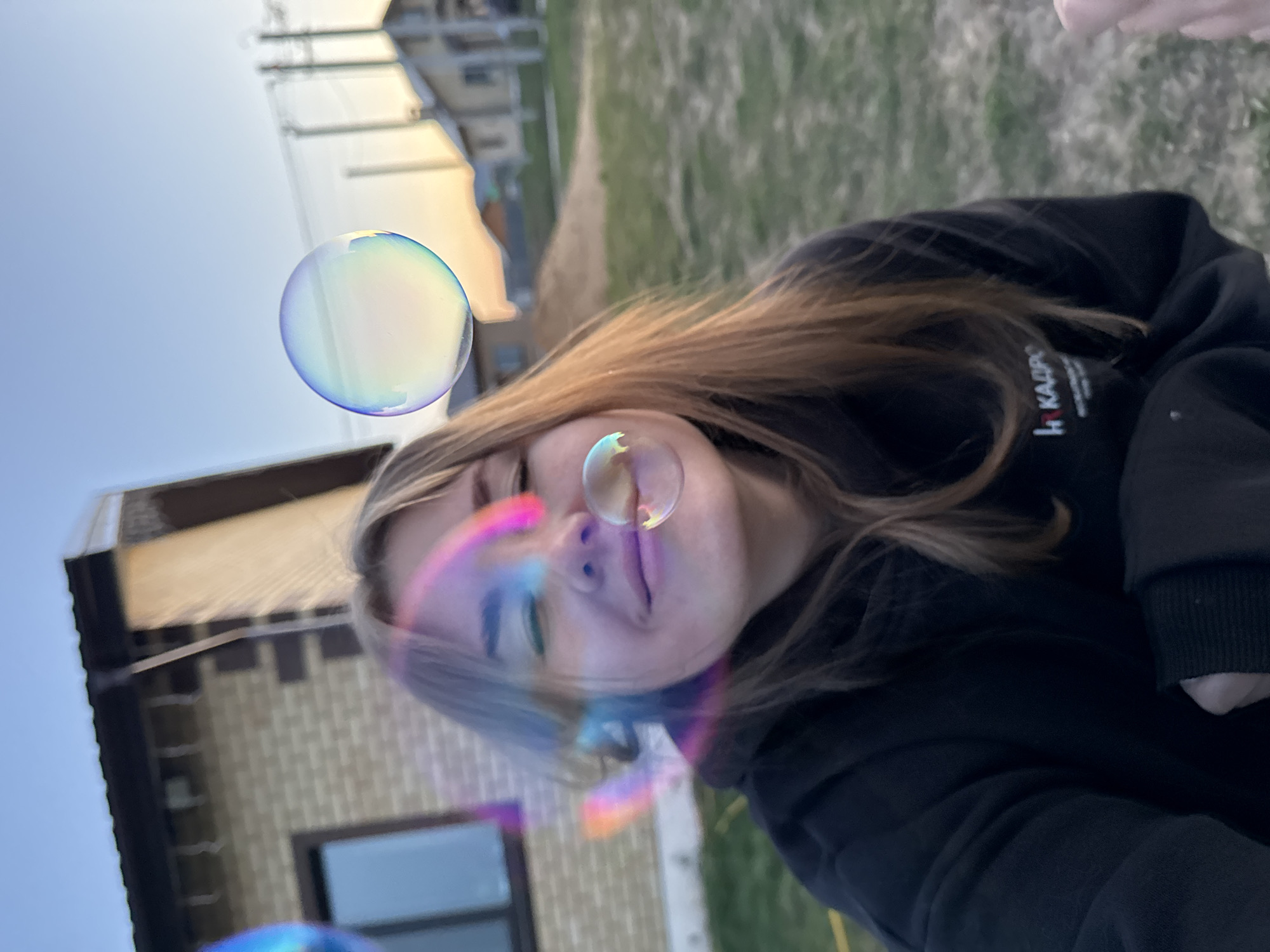 The image shows a selfie of a young woman with fair hair wearing a dark sweatshirt with a logo on the chest. She is partially obscured by soap bubbles of varying sizes. The background includes a building with a light-colored brick exterior and a dark window, and a patch of grass leading to a distant building under a clear sky. The location is identified as Ферма, Russia based on geotagging information.