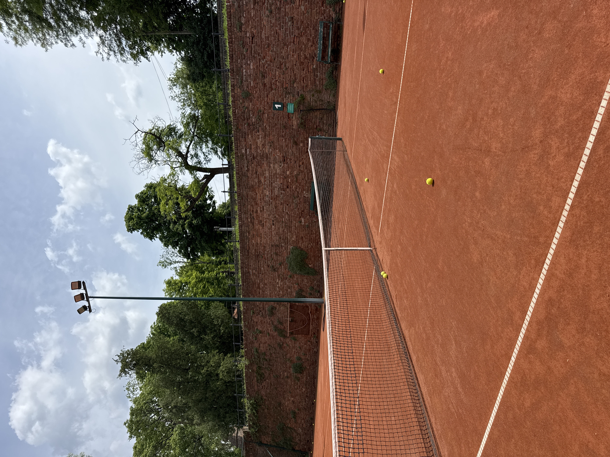 The image shows a red clay tennis court in Belgrade, Serbia. The court is surrounded by a high brick wall, trees, and a tall light post. Four tennis balls are scattered on the court surface. A green sign with the number "1" is affixed to the brick wall. A tennis net divides the court. A bench is visible on the wall. The sky is partly cloudy.